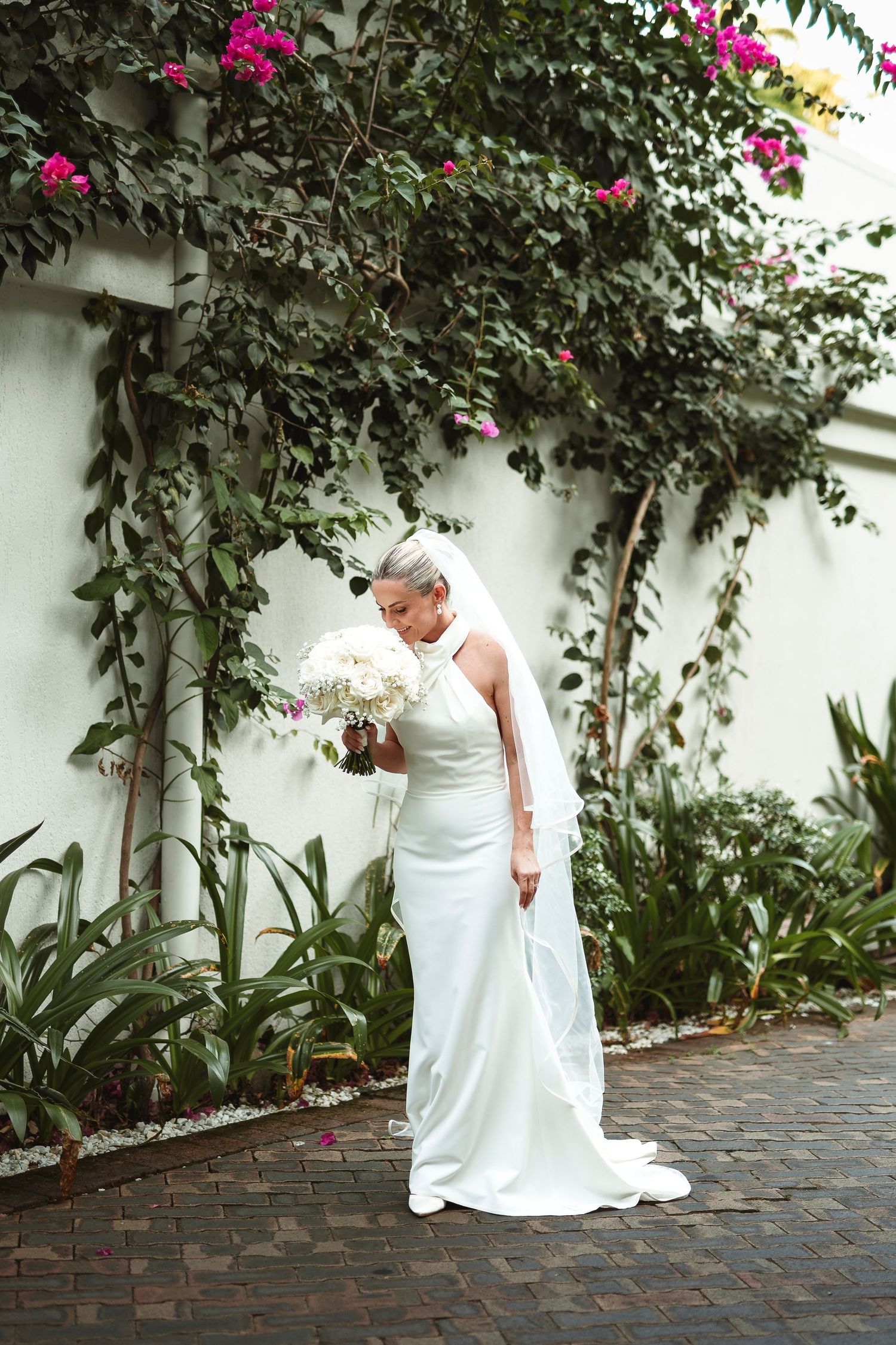 Bride in elegant white halter gown holding bouquet, standing before lush green vines and pink flowers on white wall.