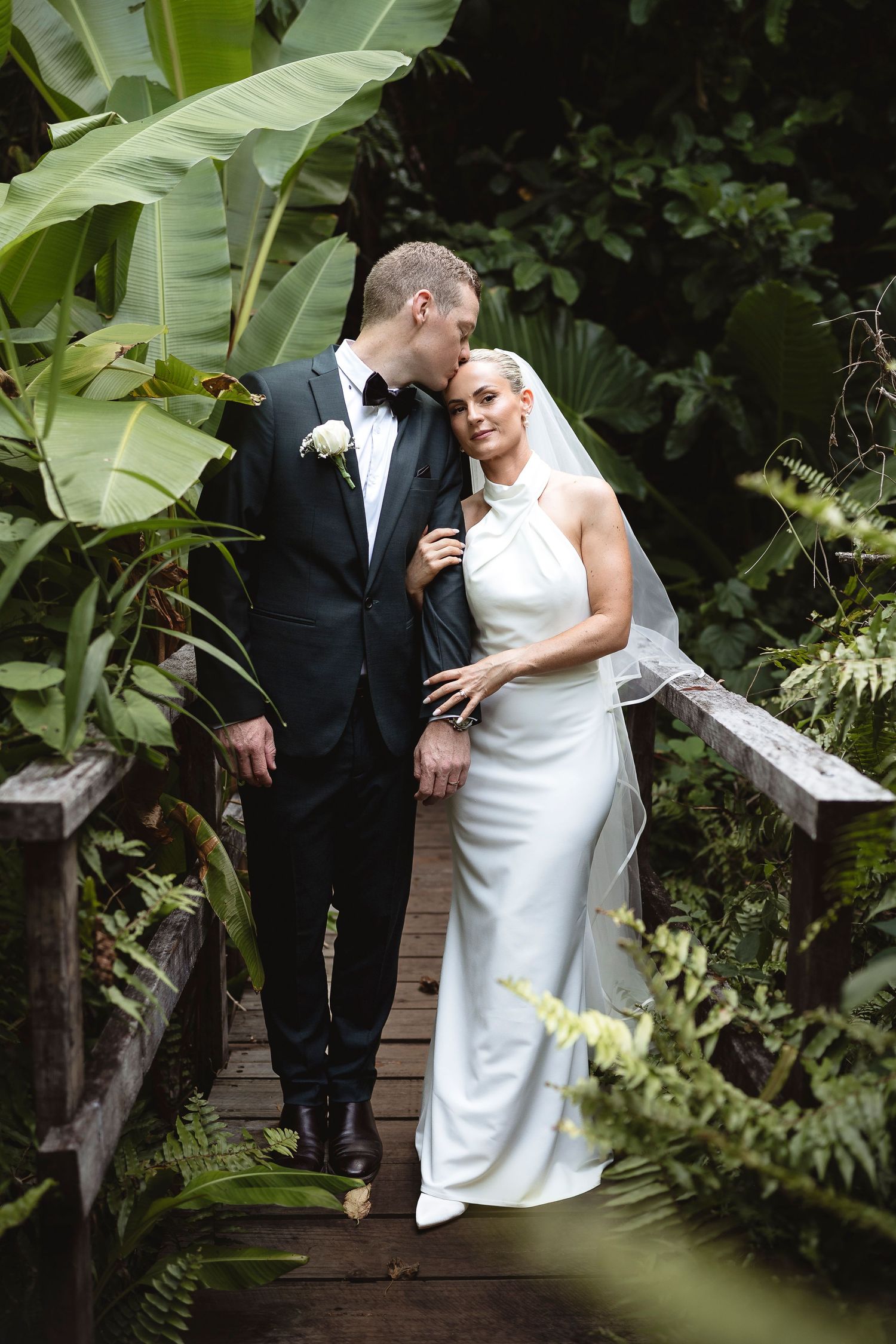 Bride and groom embrace on a wooden bridge surrounded by lush tropical greenery, bride in white halter gown with veil.