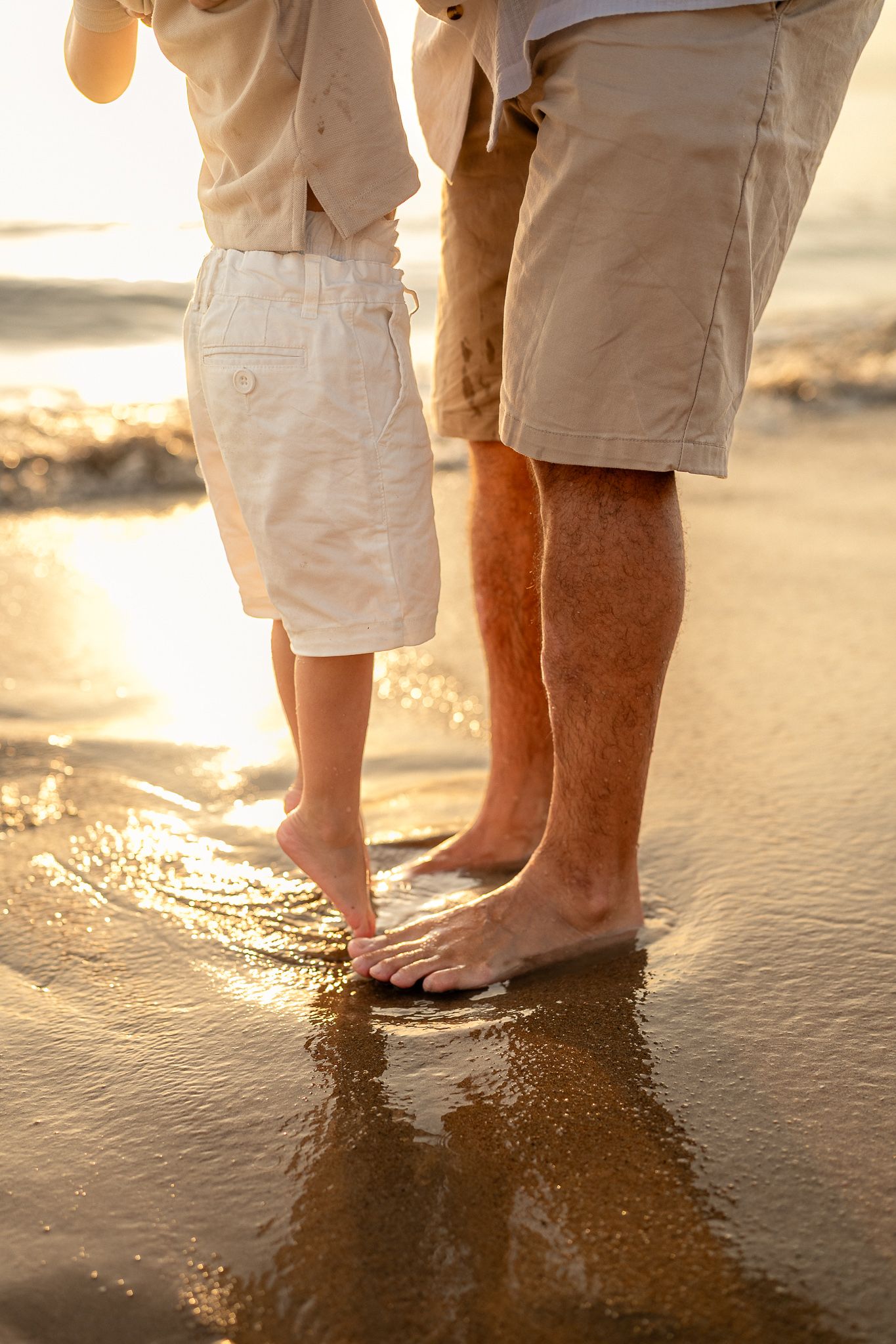 Close-up of adult and child standing barefoot together in ocean waves on a golden sunset beach.