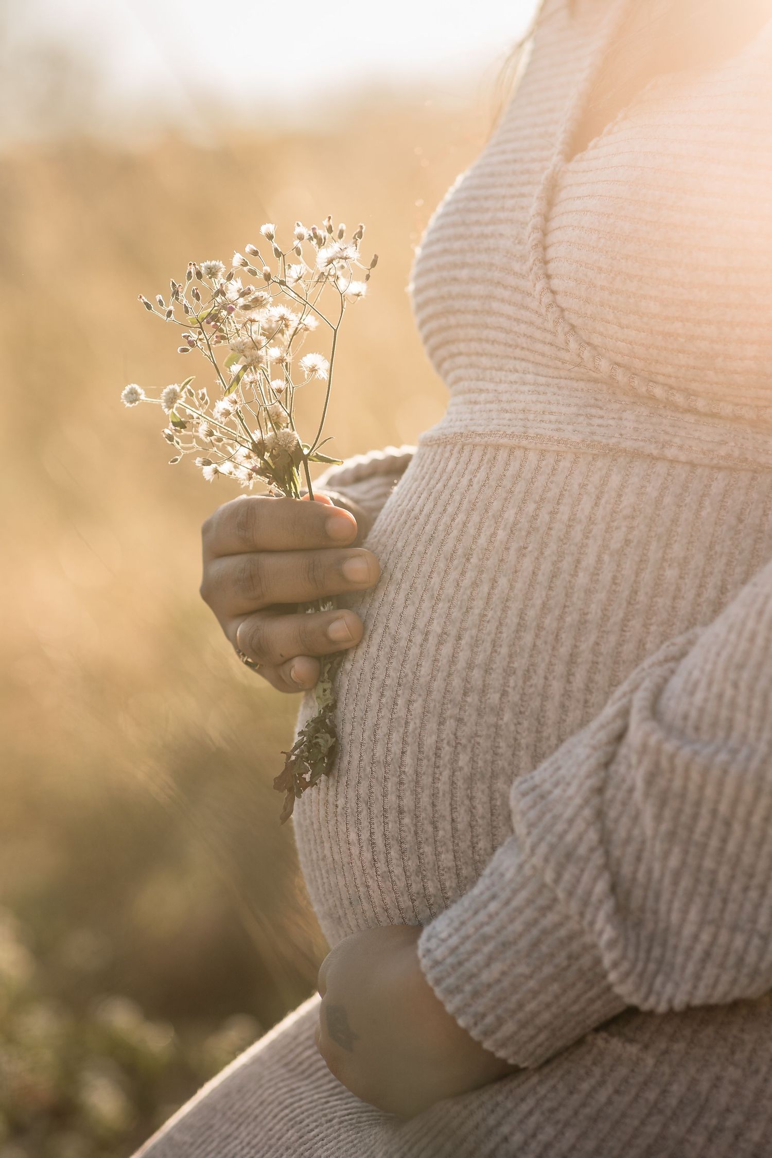 Pregnant woman in beige knit sweater holds wildflowers in golden sunlight outdoors.