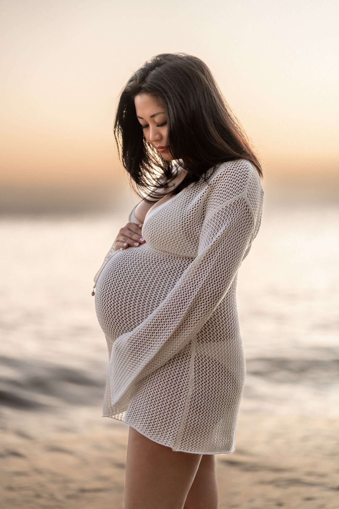 Pregnant woman in white mesh cover-up cradling her baby bump on a beach at sunset.