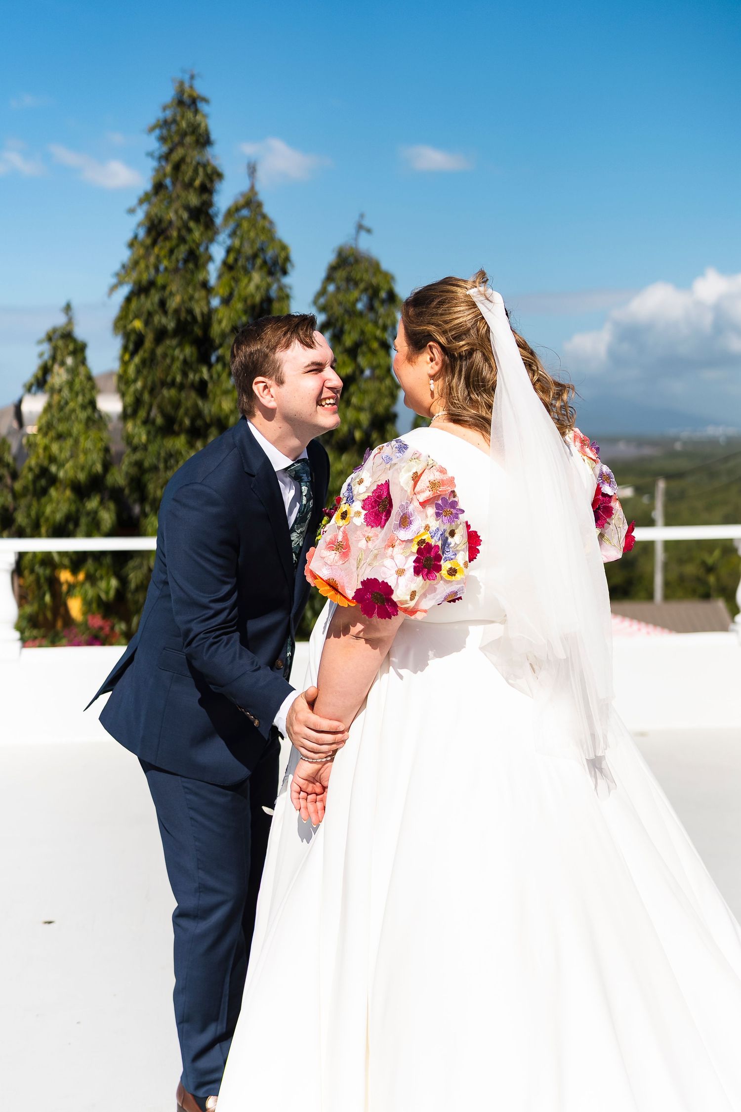 Bride in floral-sleeved gown and groom in navy suit laughing together on rooftop with trees and blue sky.