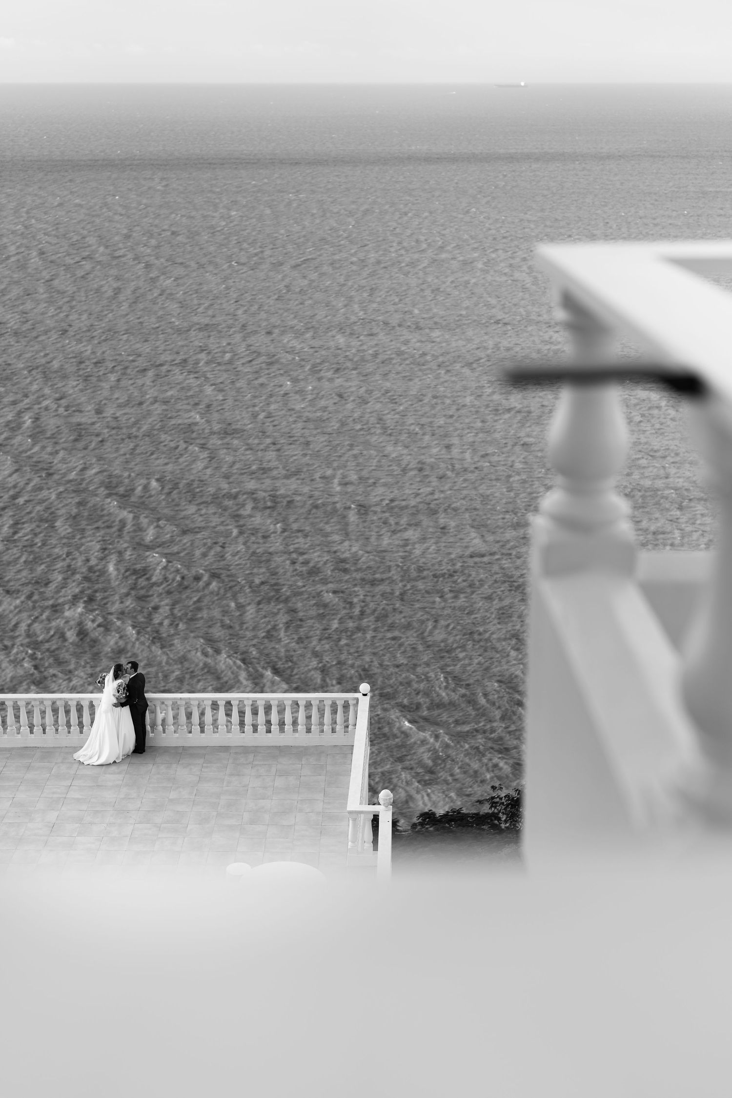 Black and white photo of a couple embracing on a seaside terrace with ocean backdrop.
