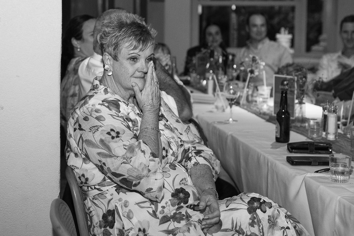 Emotional woman in floral dress with hands clasped at formal dinner reception in black and white photo.