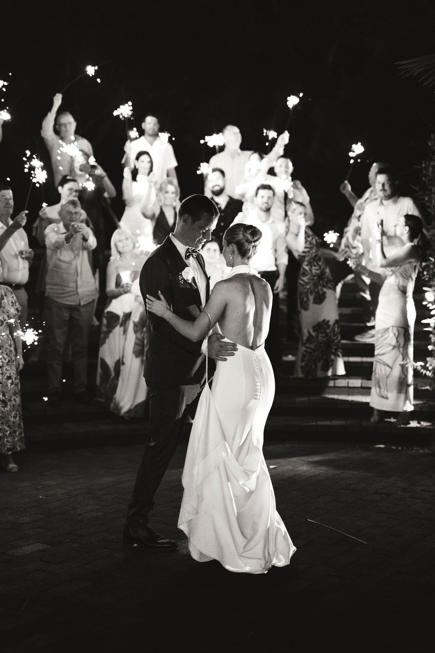 Black and white photo of bride and groom sharing first dance surrounded by guests holding sparklers at wedding reception.