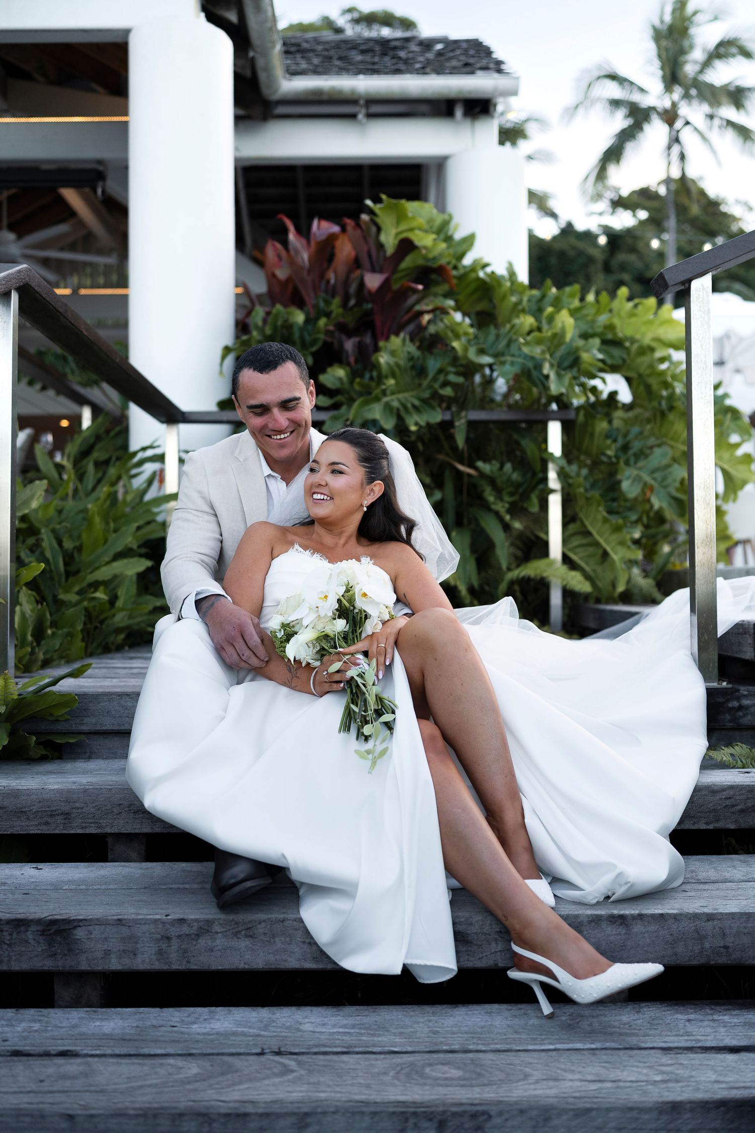 A smiling bride and groom sit on wooden steps surrounded by tropical greenery, she holds a white bouquet in her wedding dress.