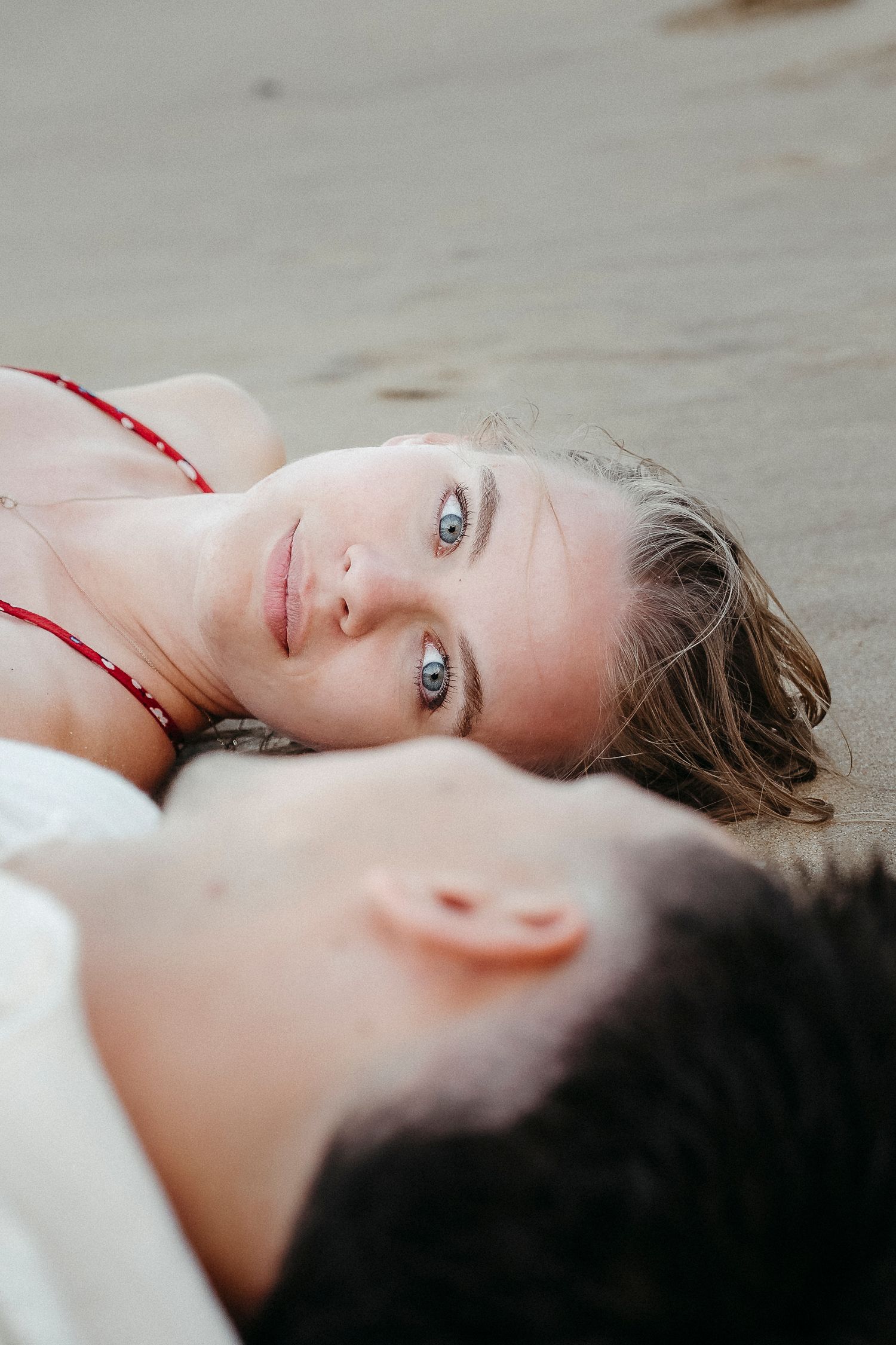 A young woman with blue eyes lies on sandy beach beside a dark-haired man, gazing softly at the camera.