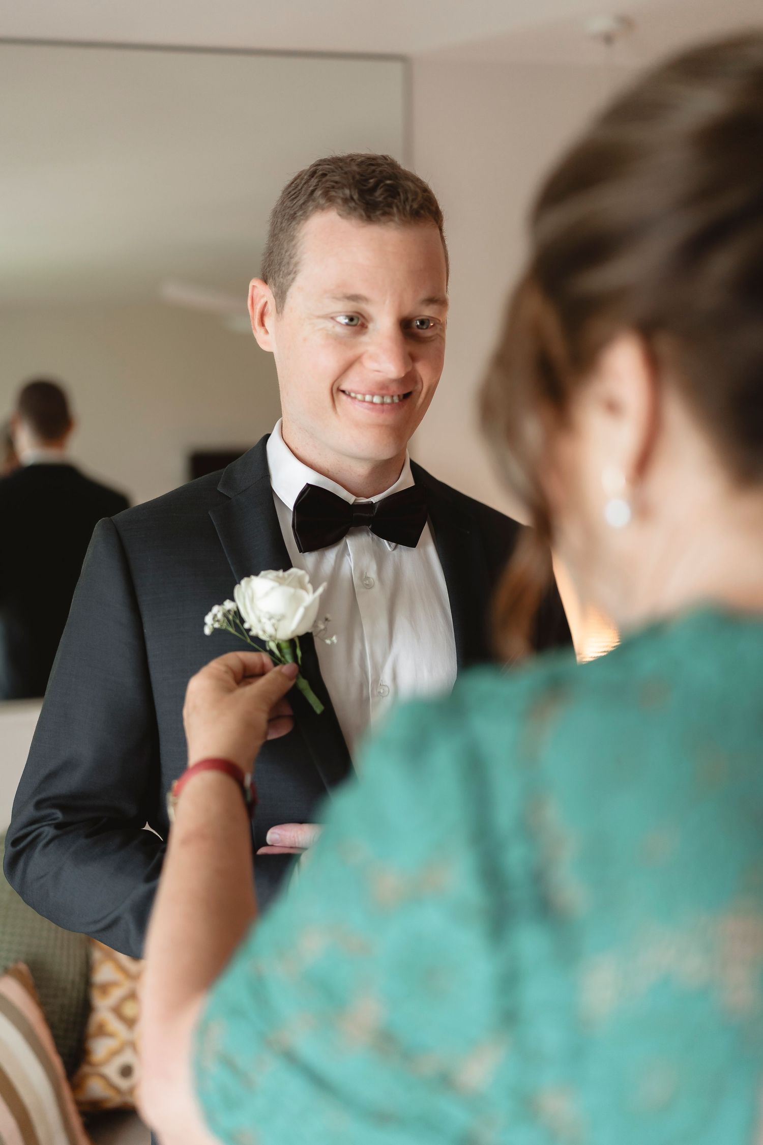 A smiling groom in a tuxedo gets a white boutonniere pinned by a woman in a teal dress before the wedding.