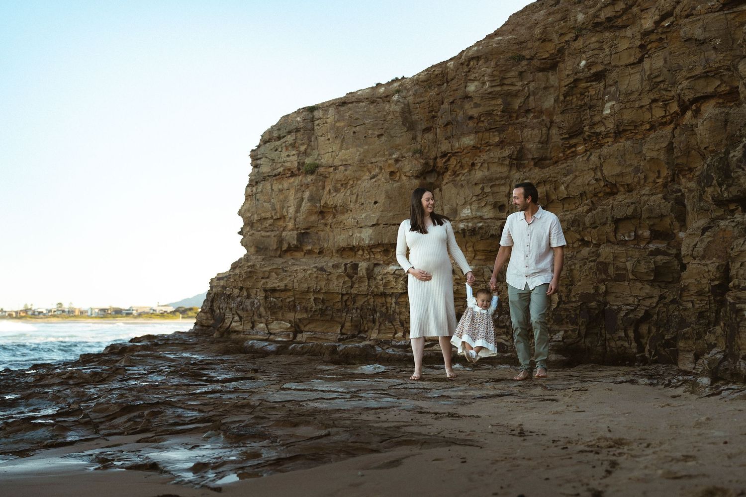 Pregnant woman in white dress with partner and child walking on rocky beach shore at sunset.