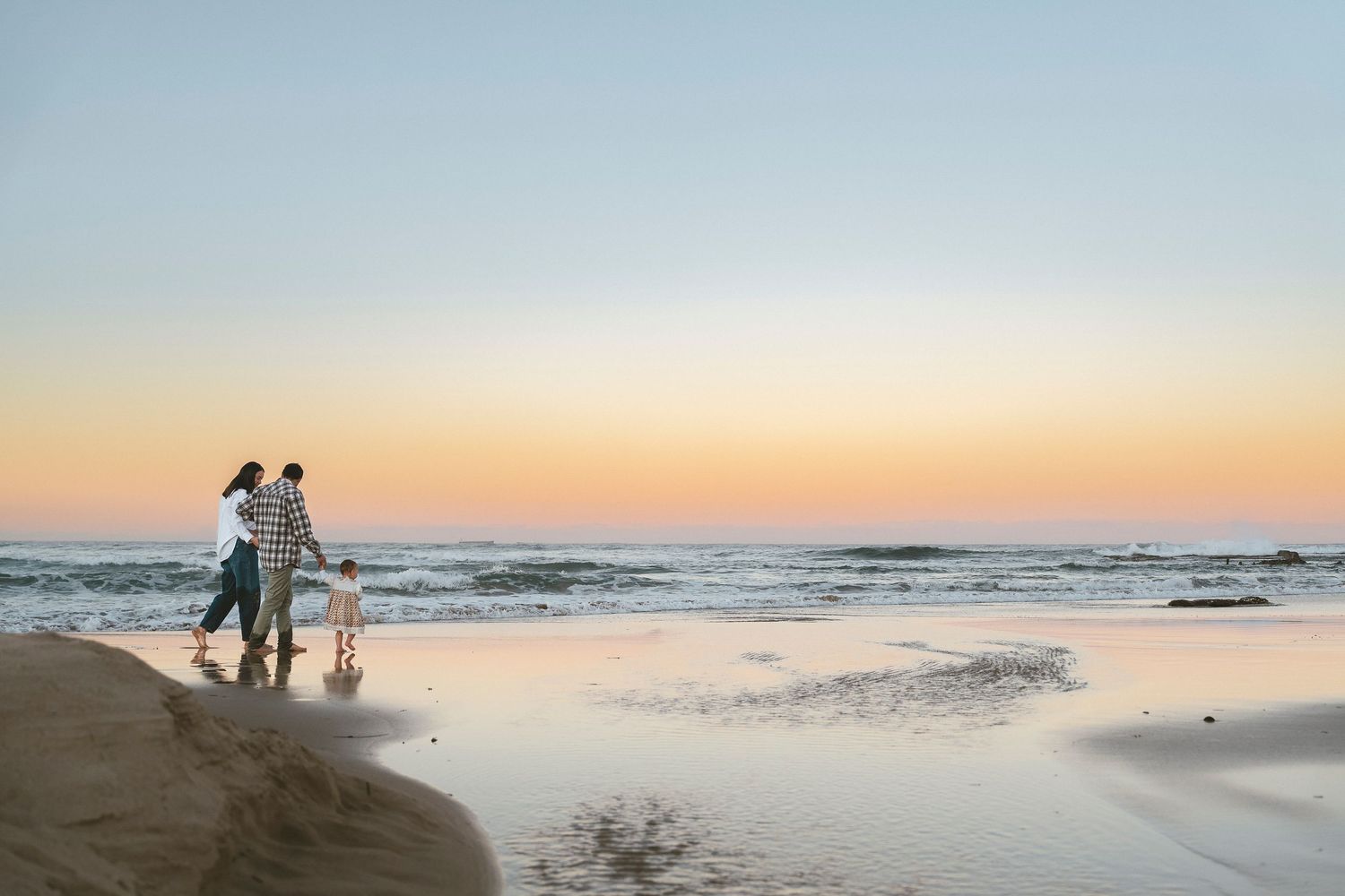 Family of three walking on a sandy beach at sunset, waves gently crashing as golden light reflects on the shore.