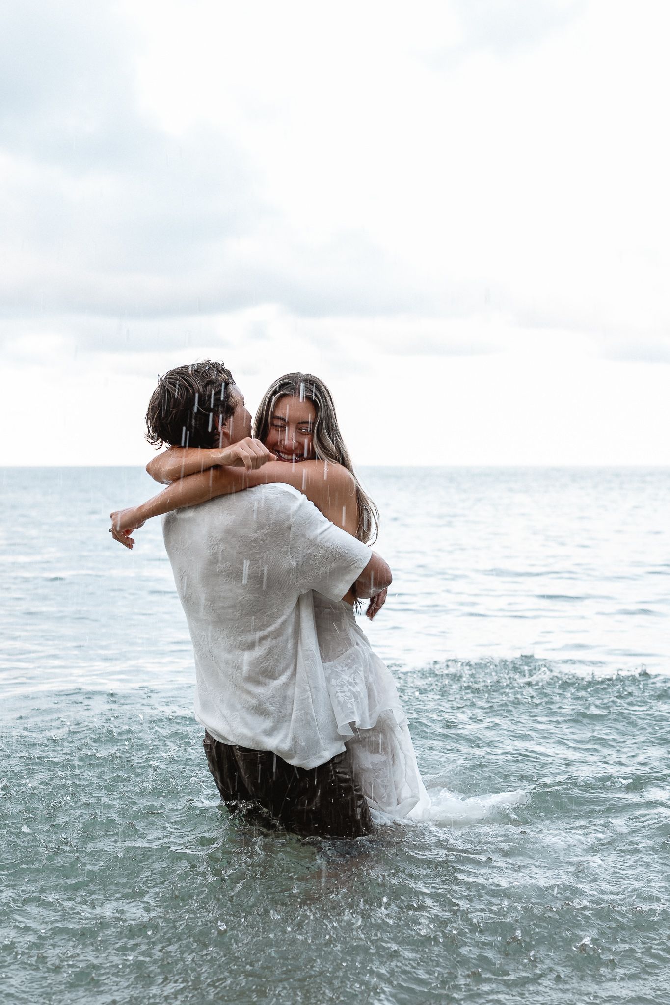 Couple embracing in ocean waves fully clothed in white during a romantic beach shoot.
