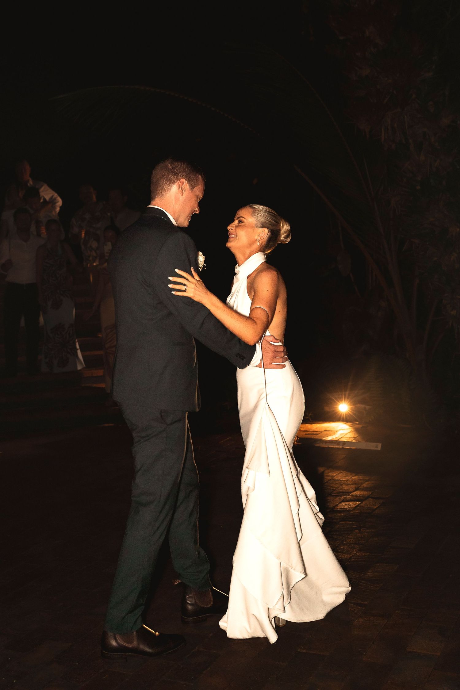 Bride and groom share first dance outdoors at night, bride in elegant backless white gown, groom in dark suit.
