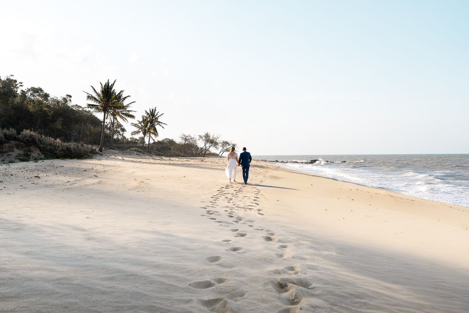 Newlywed couple walking along a pristine sandy beach lined with palm trees at sunset.