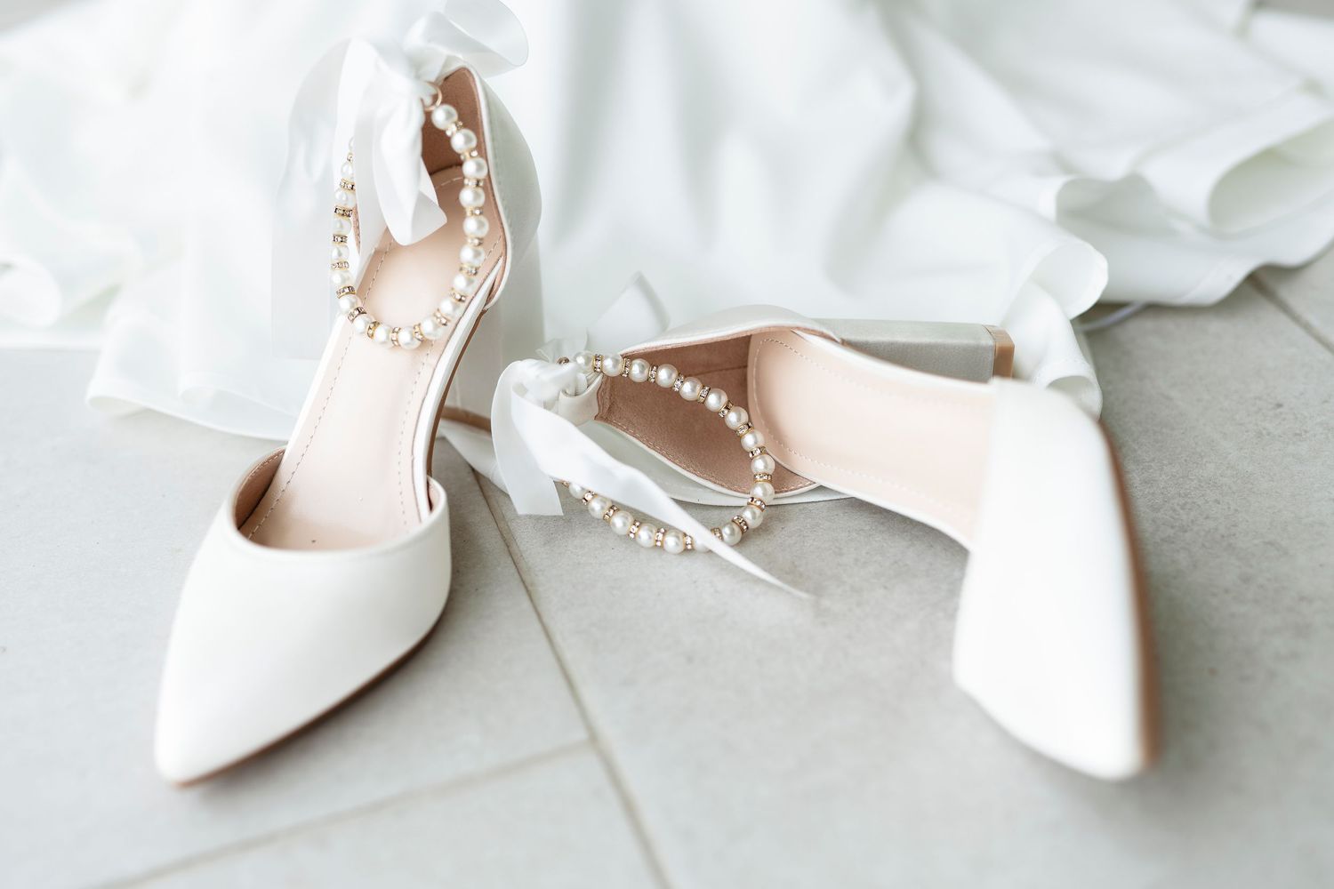 Elegant white bridal heels with pearl ankle straps and ribbon bows on marble floor.
