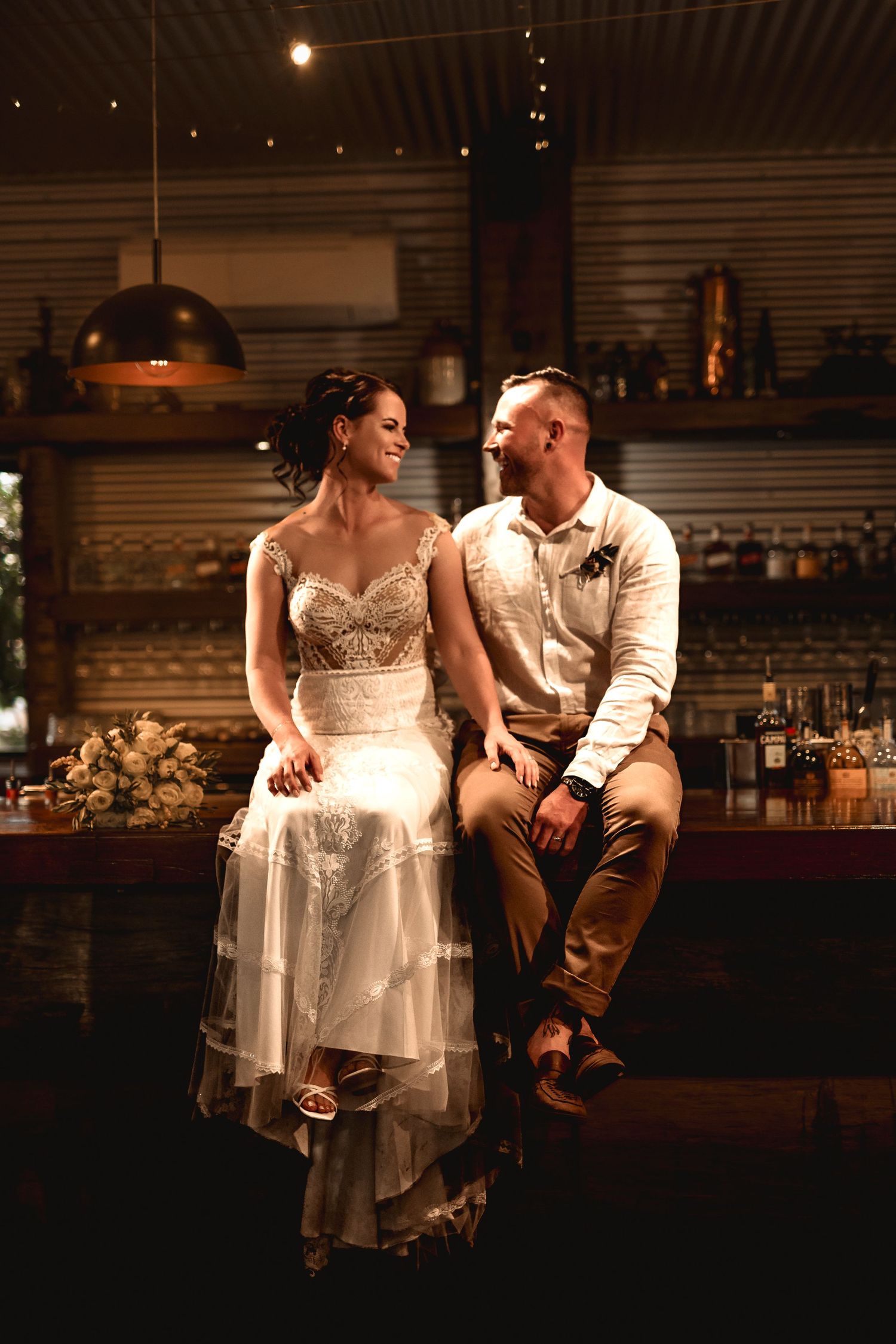 Bride and groom sitting together at a warmly lit rustic bar, sharing a romantic glance in wedding attire.