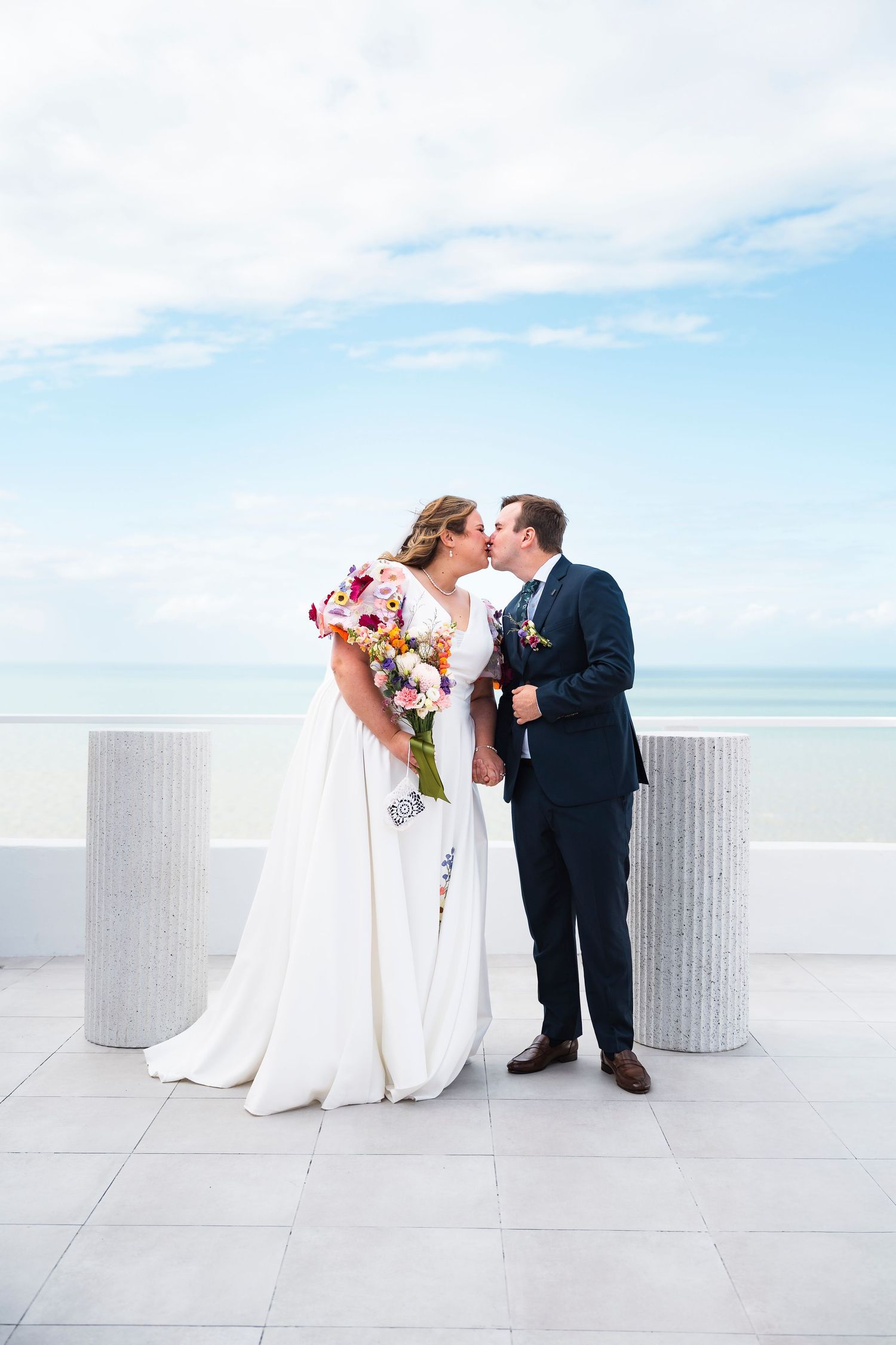 Bride and groom share a kiss on a rooftop terrace with ocean views, bride holding a colorful bouquet in a white gown.