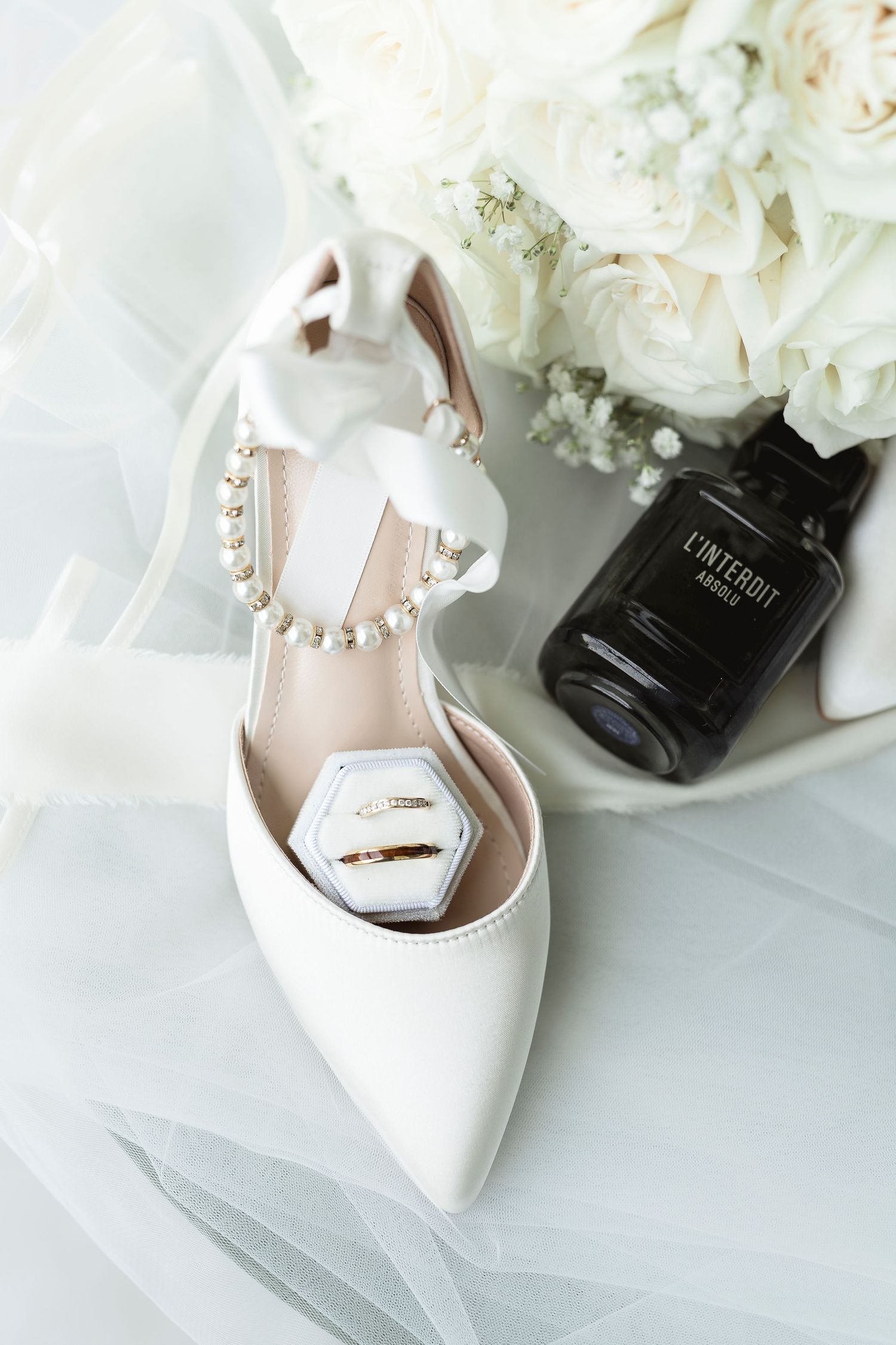 White bridal heels with pearl straps, gold wedding rings, and perfume bottle.