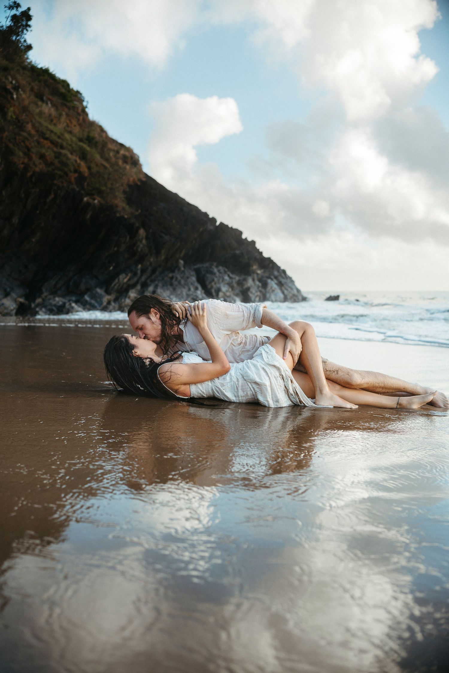 Couple sharing a romantic kiss lying on a wet sandy beach with ocean waves and rocky cliffs in the background.