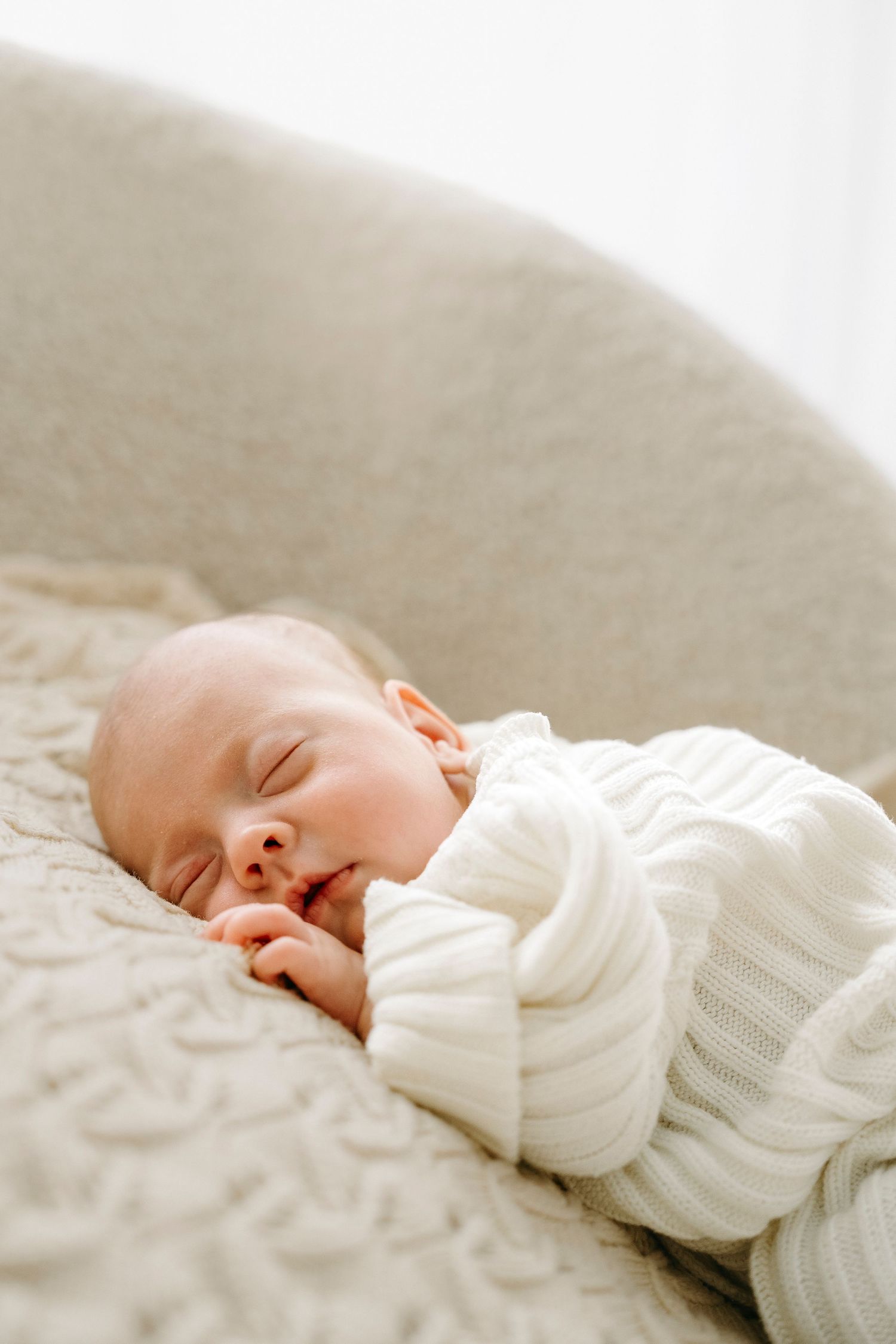 A peaceful newborn baby sleeping in a cream knit sweater on a soft beige textured blanket in a cozy chair.