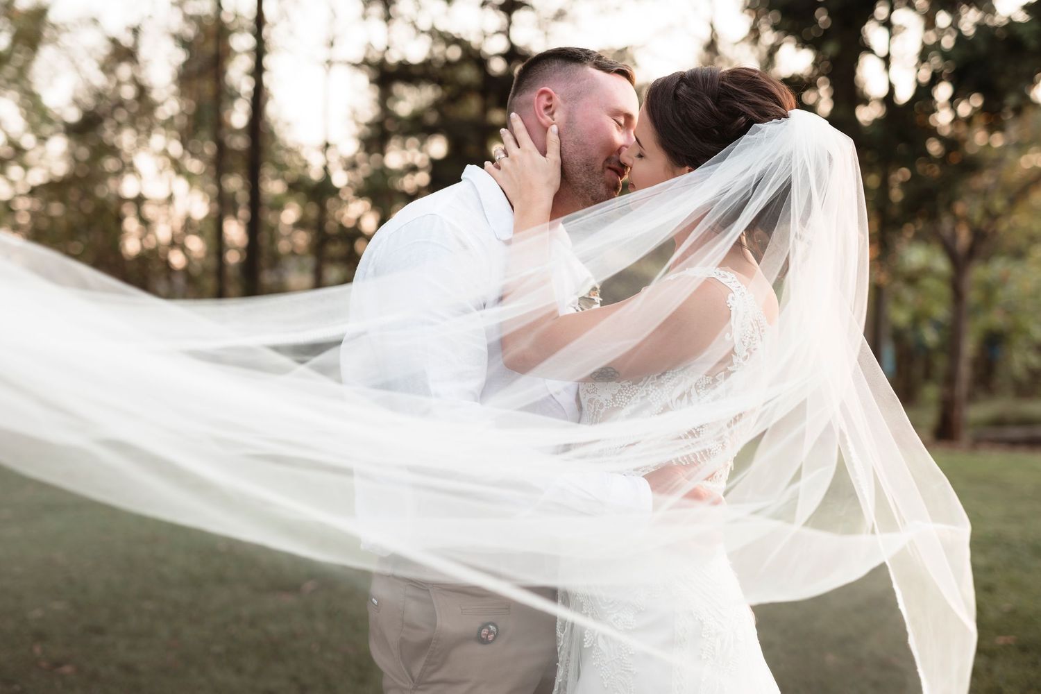 A bride and groom share a romantic kiss outdoors, with her flowing white veil billowing in the breeze.