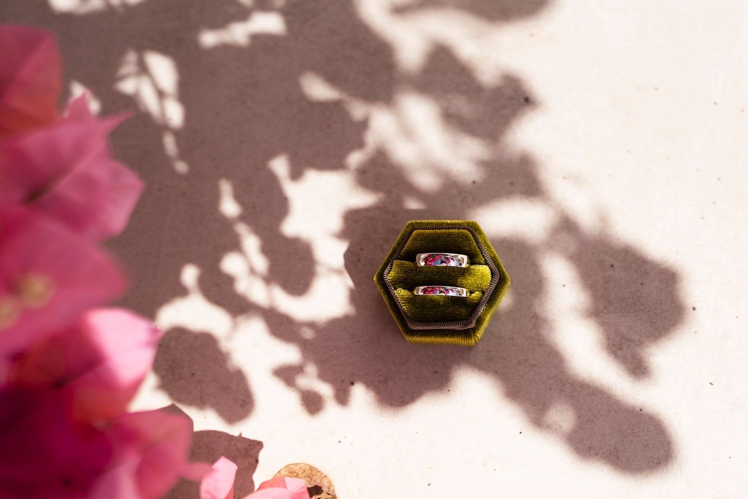 Two gold rings with pink gemstones displayed in a green hexagonal velvet box, surrounded by pink flowers and leaf shadows.