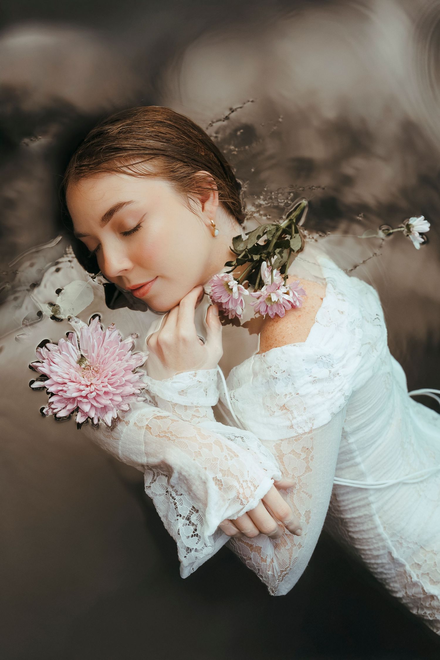 Woman in white lace dress lying peacefully with pink chrysanthemums and white flowers in dark water.