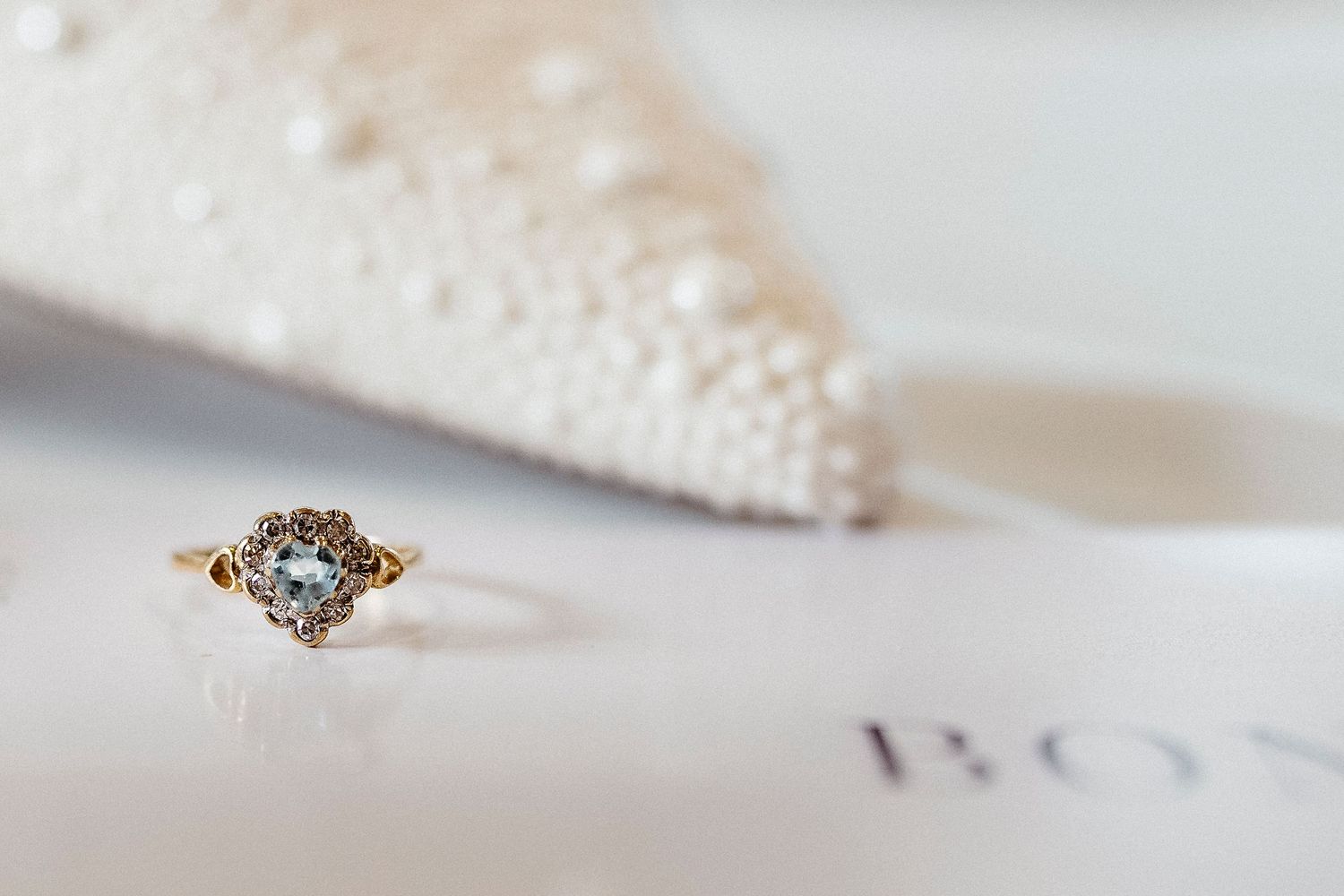 Gold halo engagement ring with center diamond displayed on white surface near pearl-adorned bridal shoe.