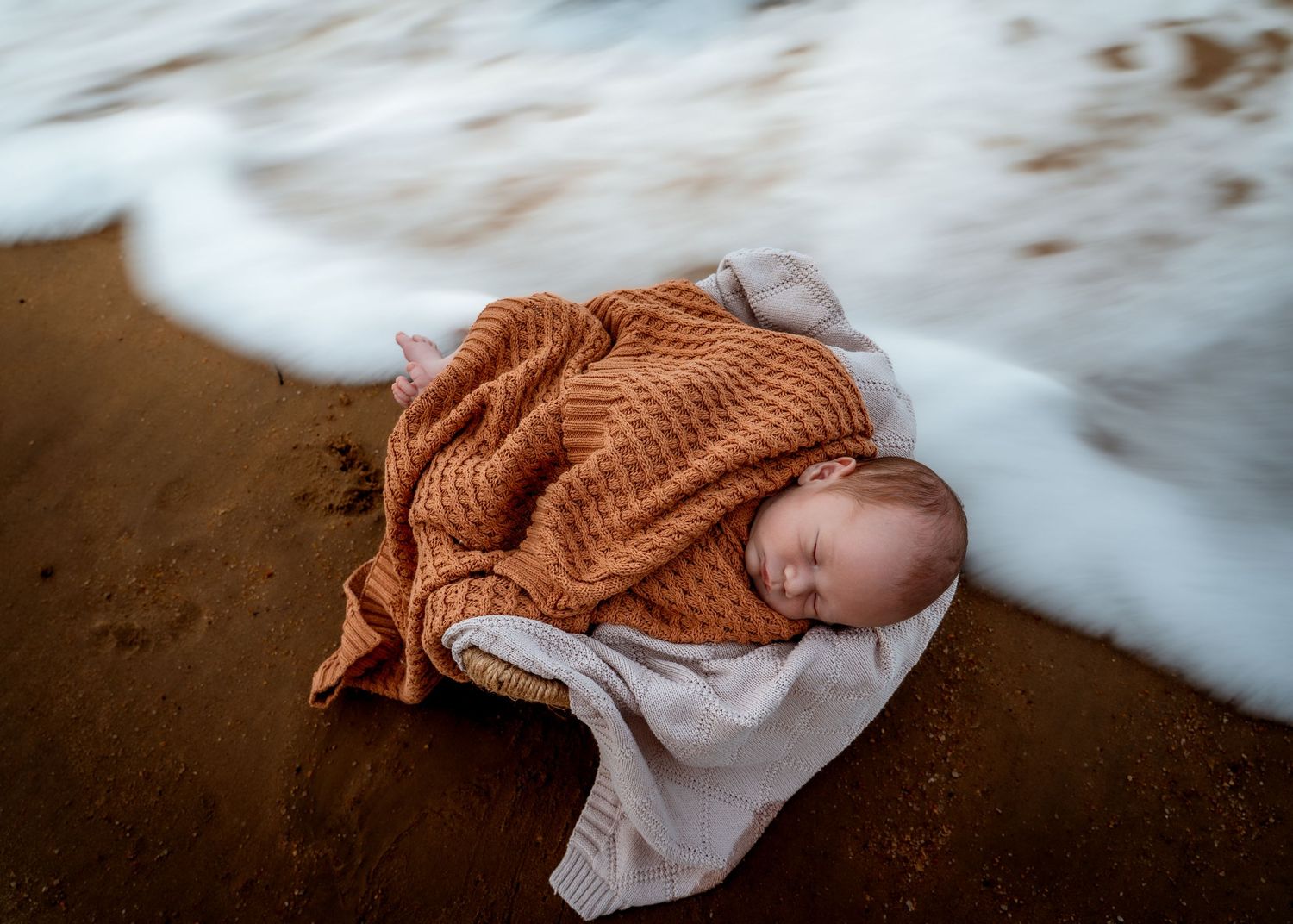 Sleeping newborn baby wrapped in orange knit blanket on sandy beach with ocean waves.