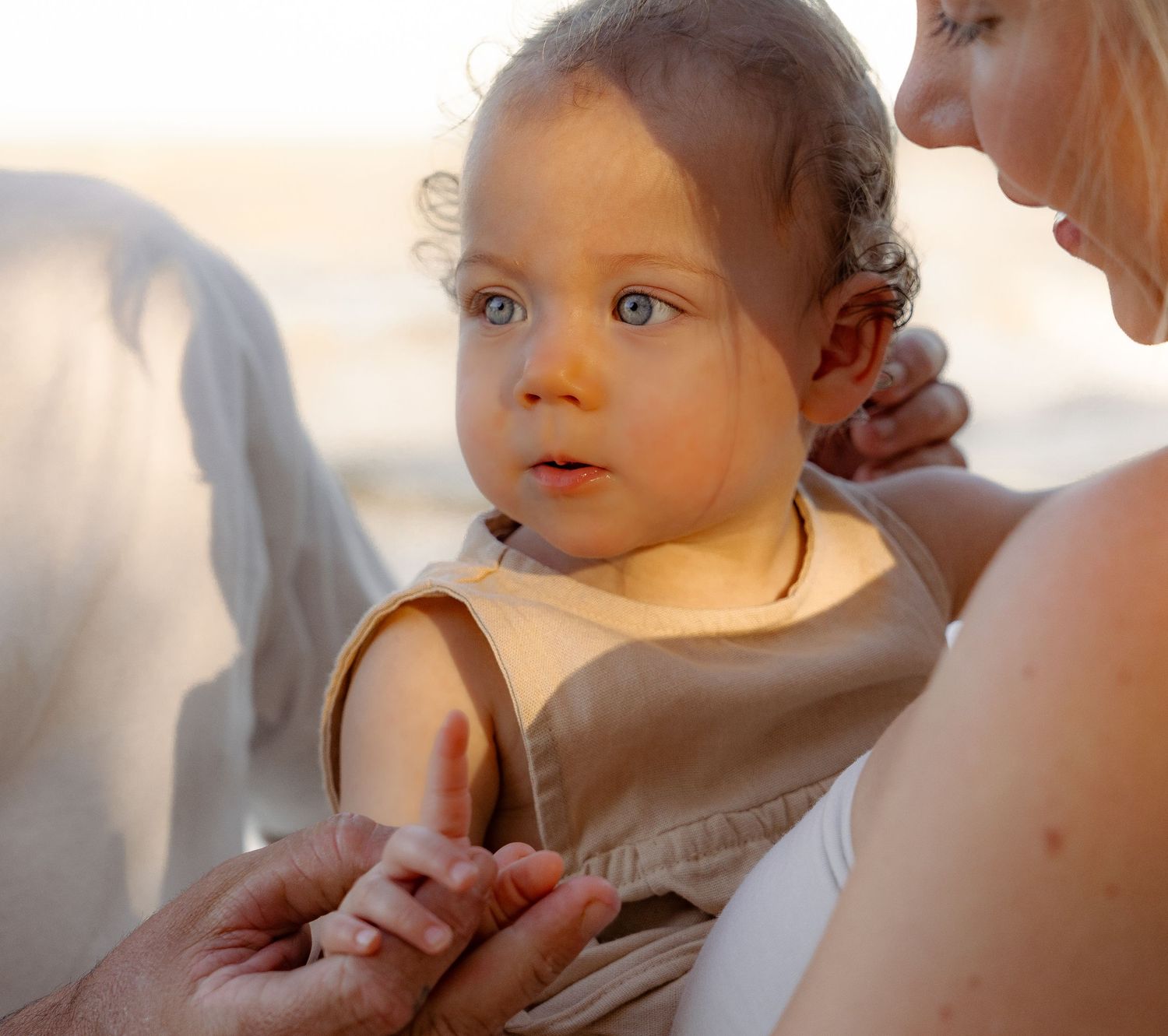 Adorable blue-eyed baby in tan romper being held by mother on a sun-kissed beach.