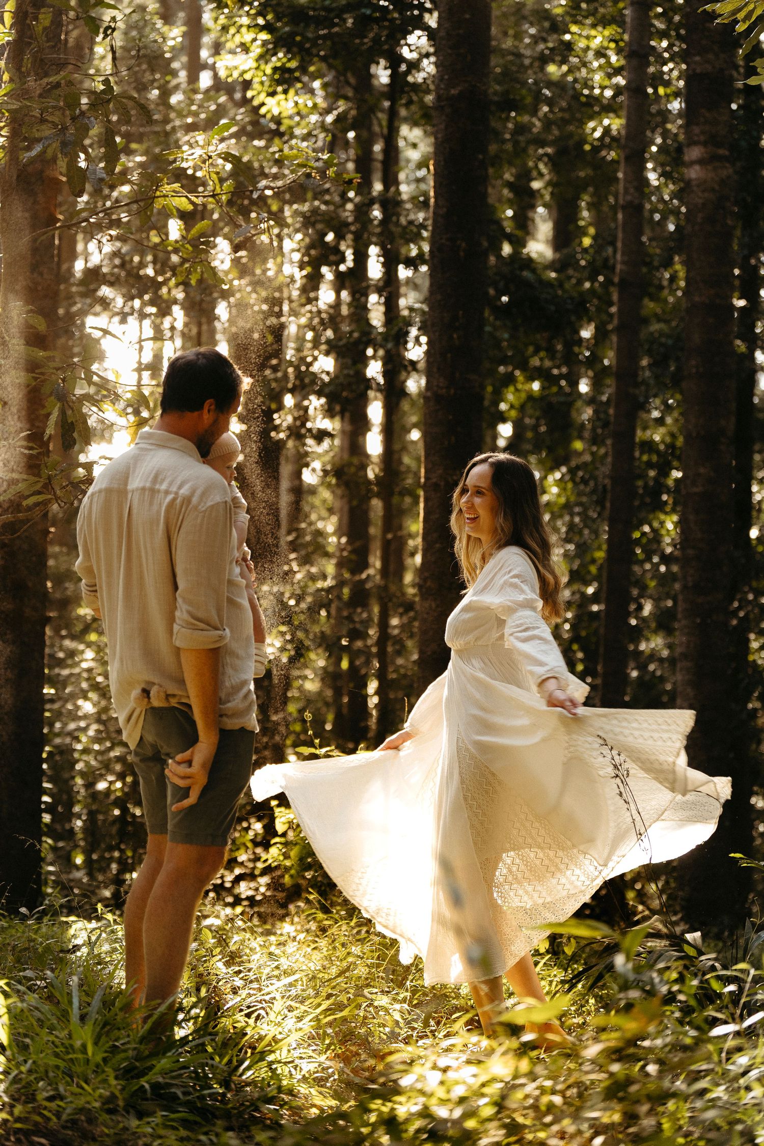 Couple dancing in sunlit forest, woman twirling white dress among tall trees in golden light.