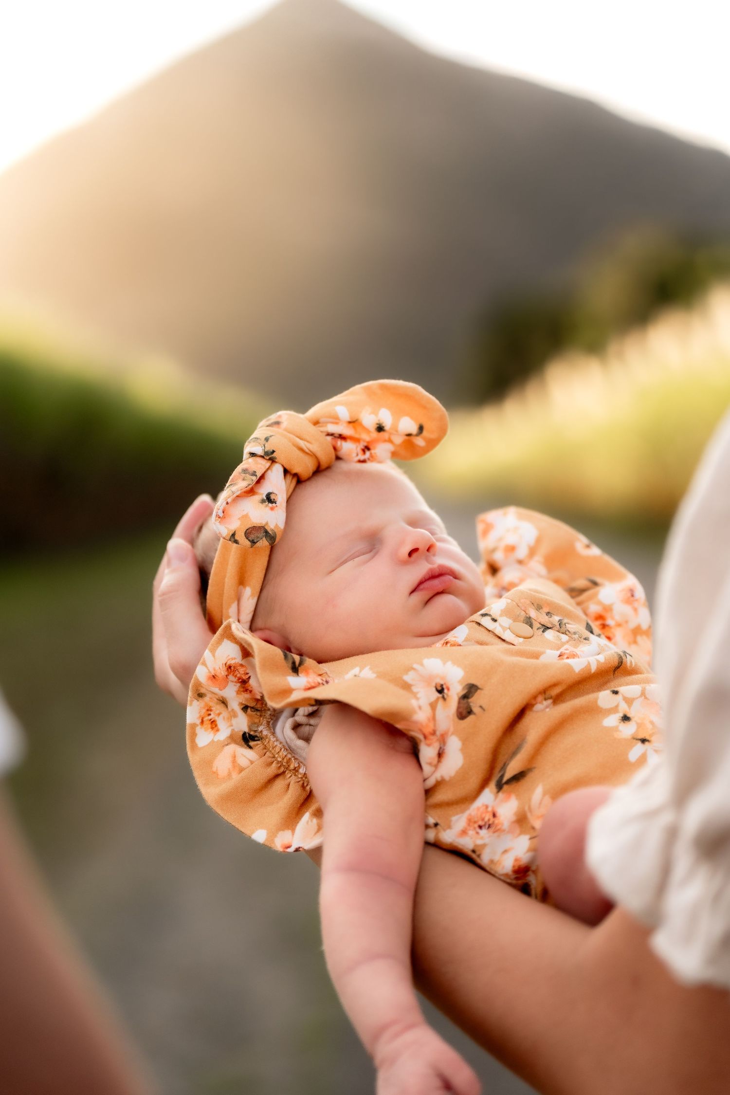 A sleeping newborn baby in an orange floral outfit held gently in arms at golden hour.