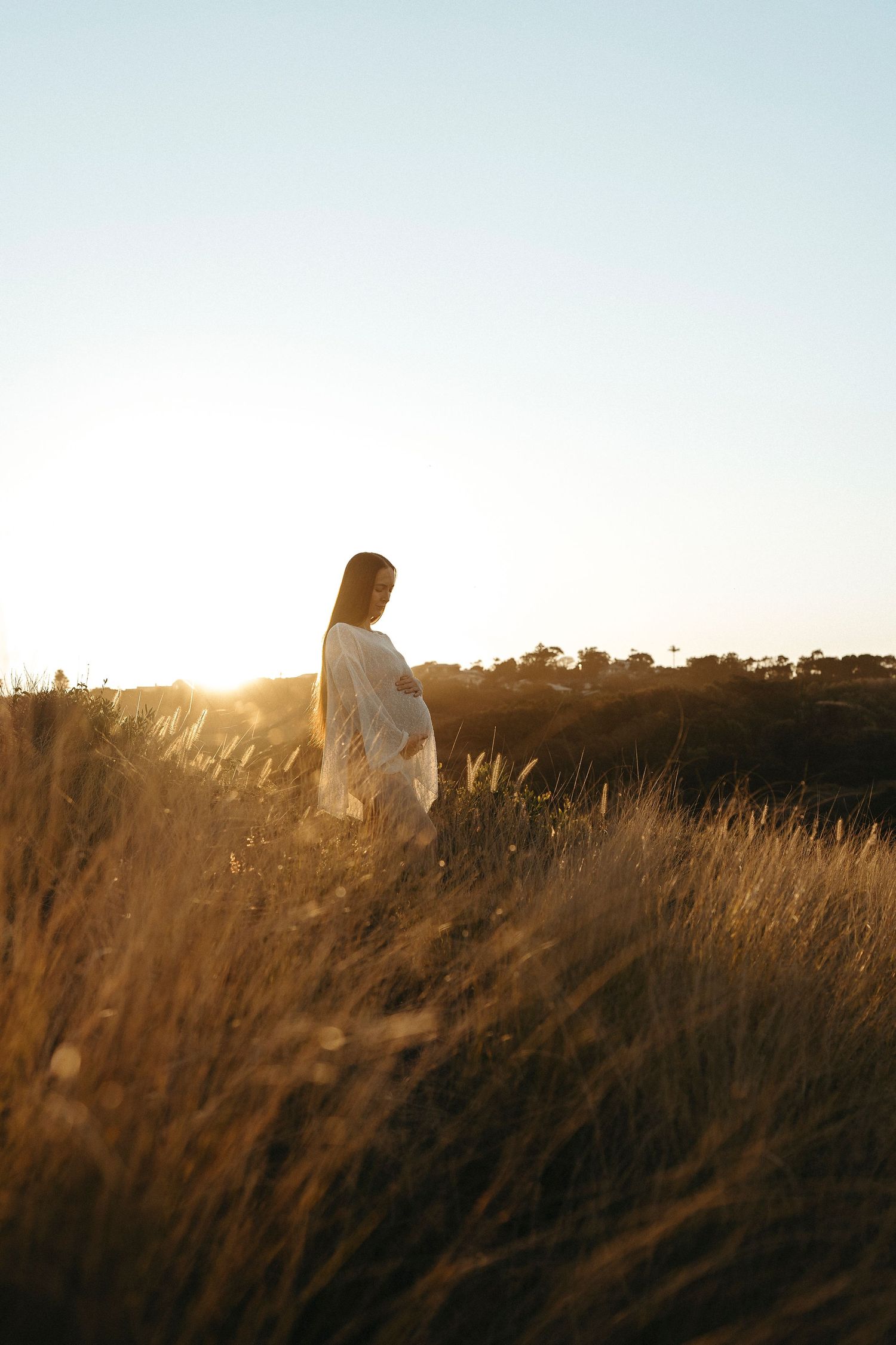 Pregnant woman in white dress standing in golden grass field at sunset, cradling her baby bump in warm sunlight.