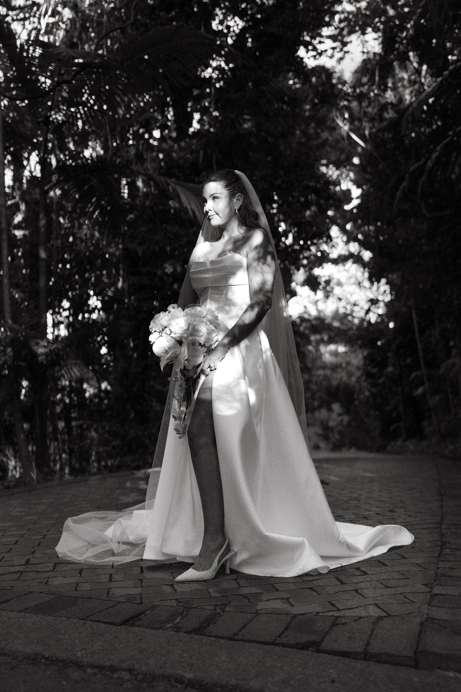 Black and white photo of a bride in a flowing gown and veil holding a bouquet, standing on a stone path surrounded by trees.