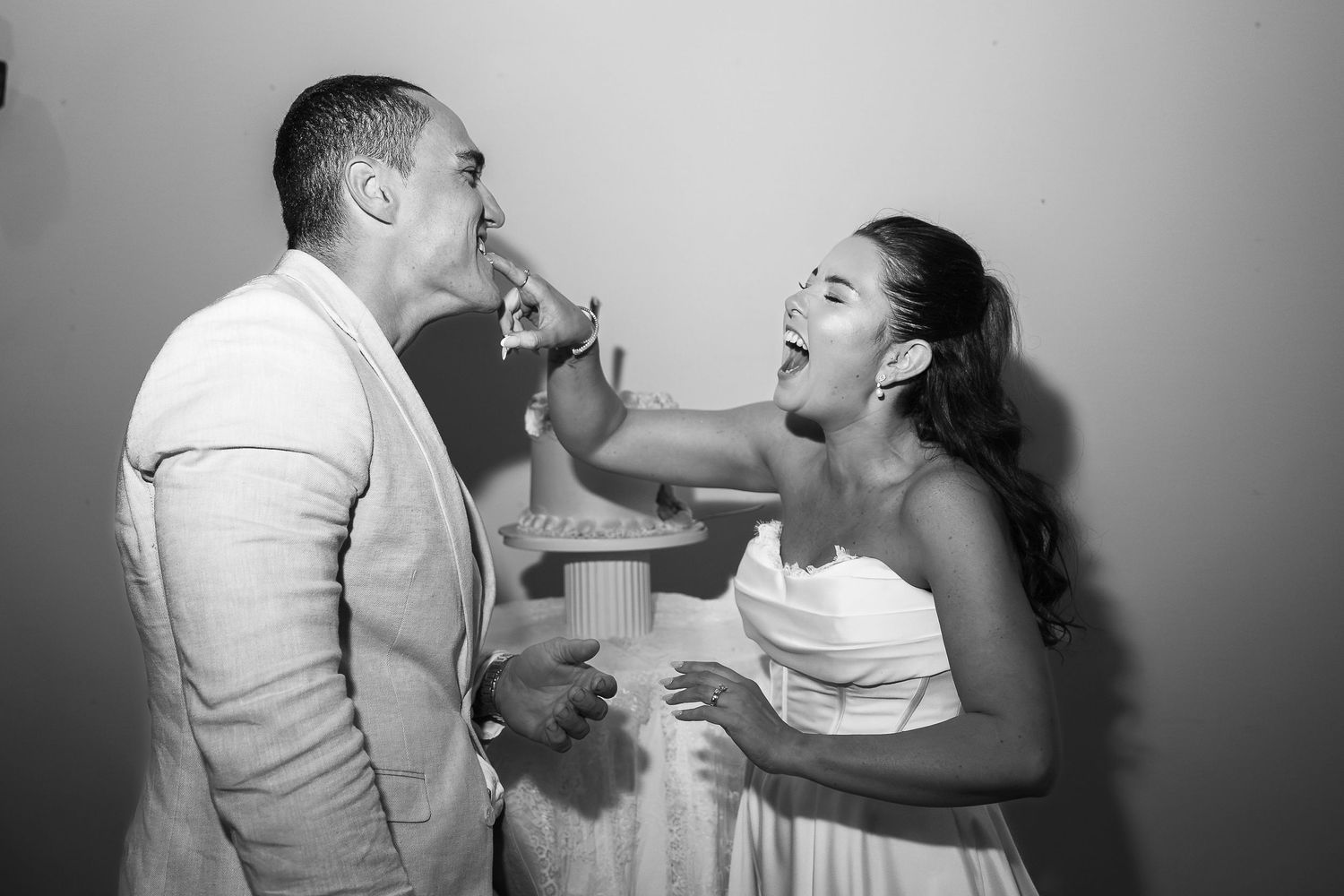Black and white photo of bride smashing wedding cake into grooms mouth at reception celebration.