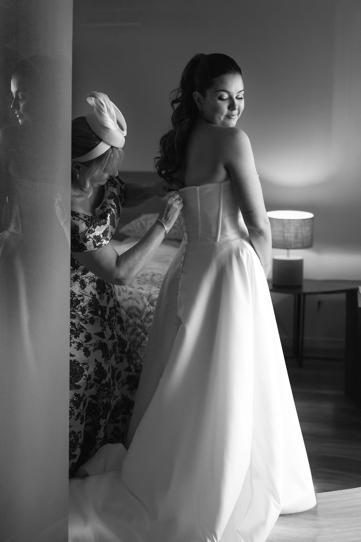Black and white photo of a bride in a white gown being helped into her dress by a woman in a floral outfit.