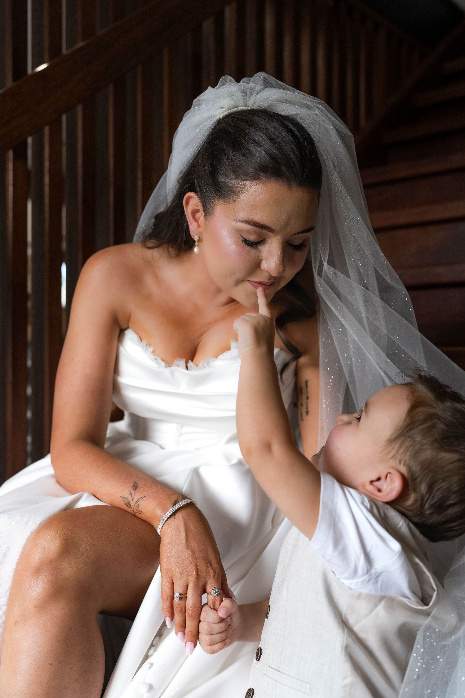 A bride in a white gown and veil shares a tender moment with a young child in white attire on her wedding day.