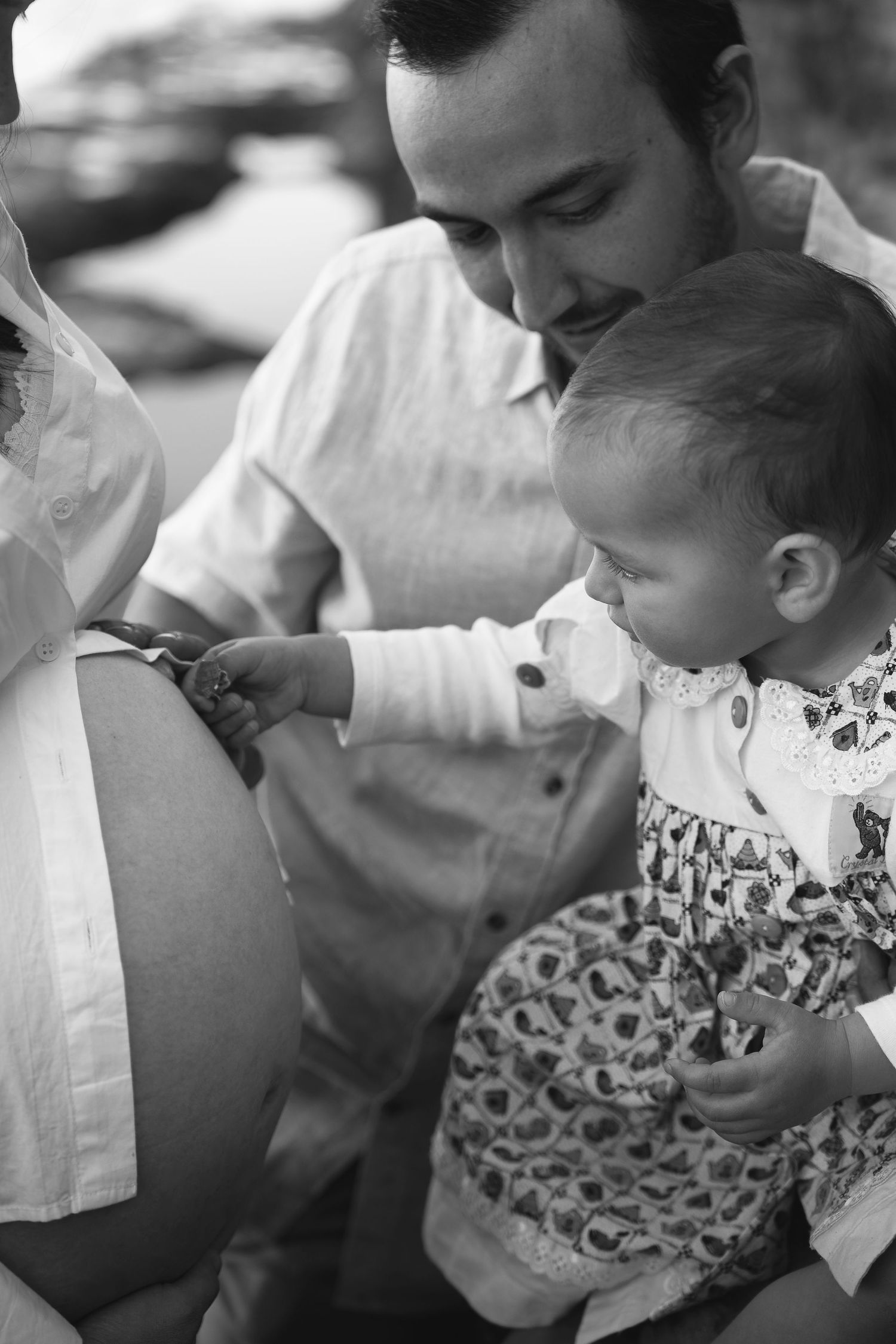 Black and white photo of a father holding a baby girl touching a pregnant woman's belly outdoors.
