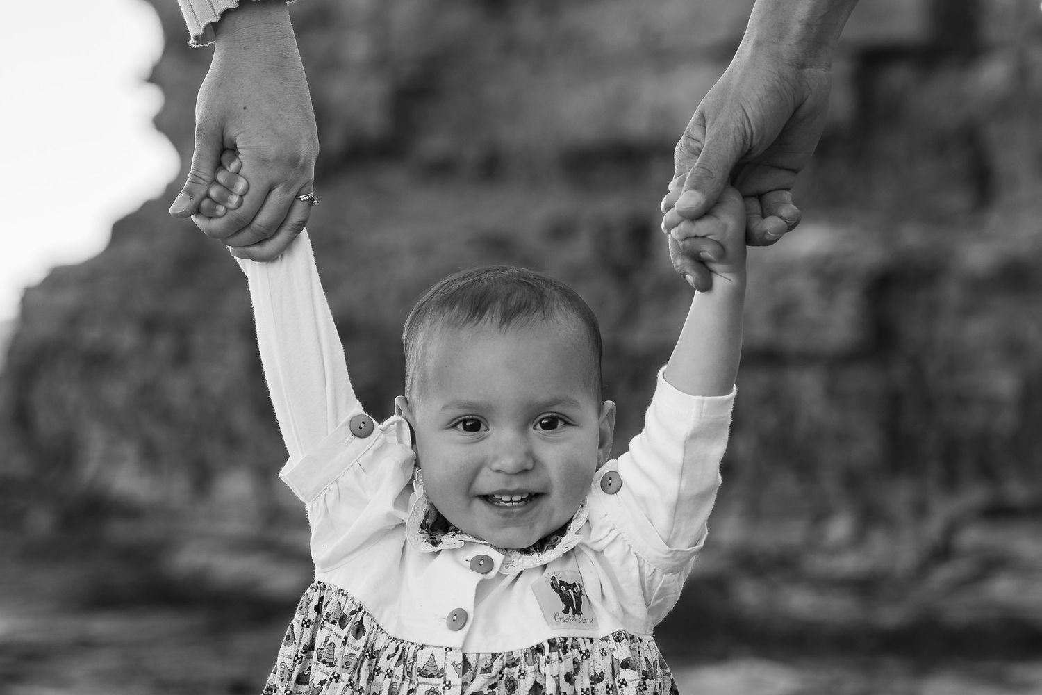 Black and white photo of a smiling baby held up by adult hands outdoors.