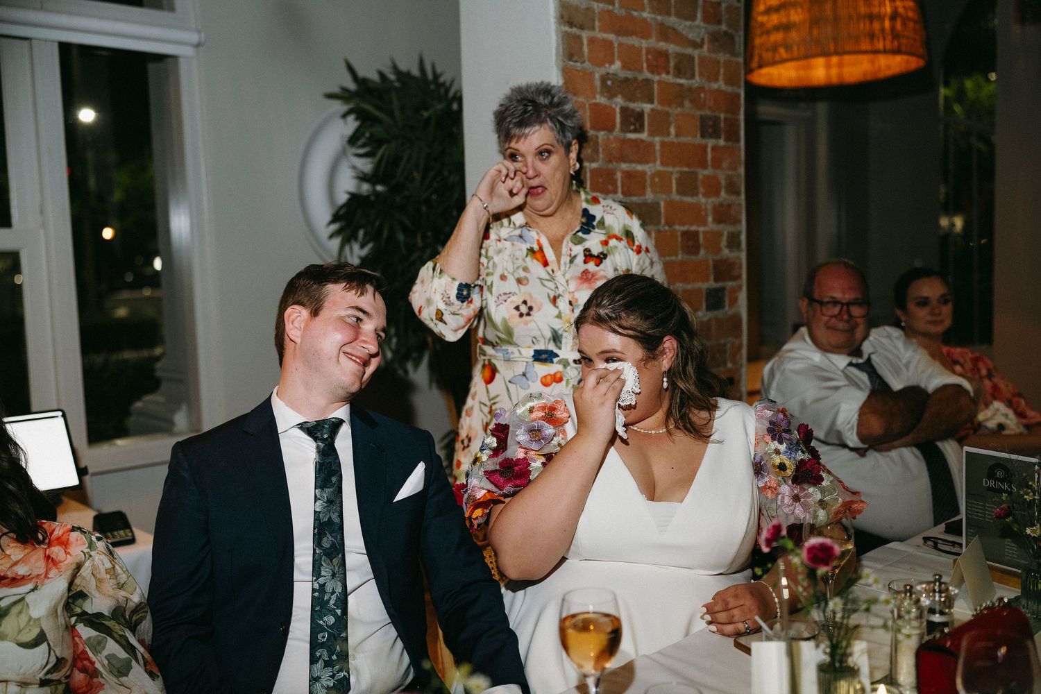 Bride and groom laugh emotionally during wedding reception speeches at elegant brick-walled venue.