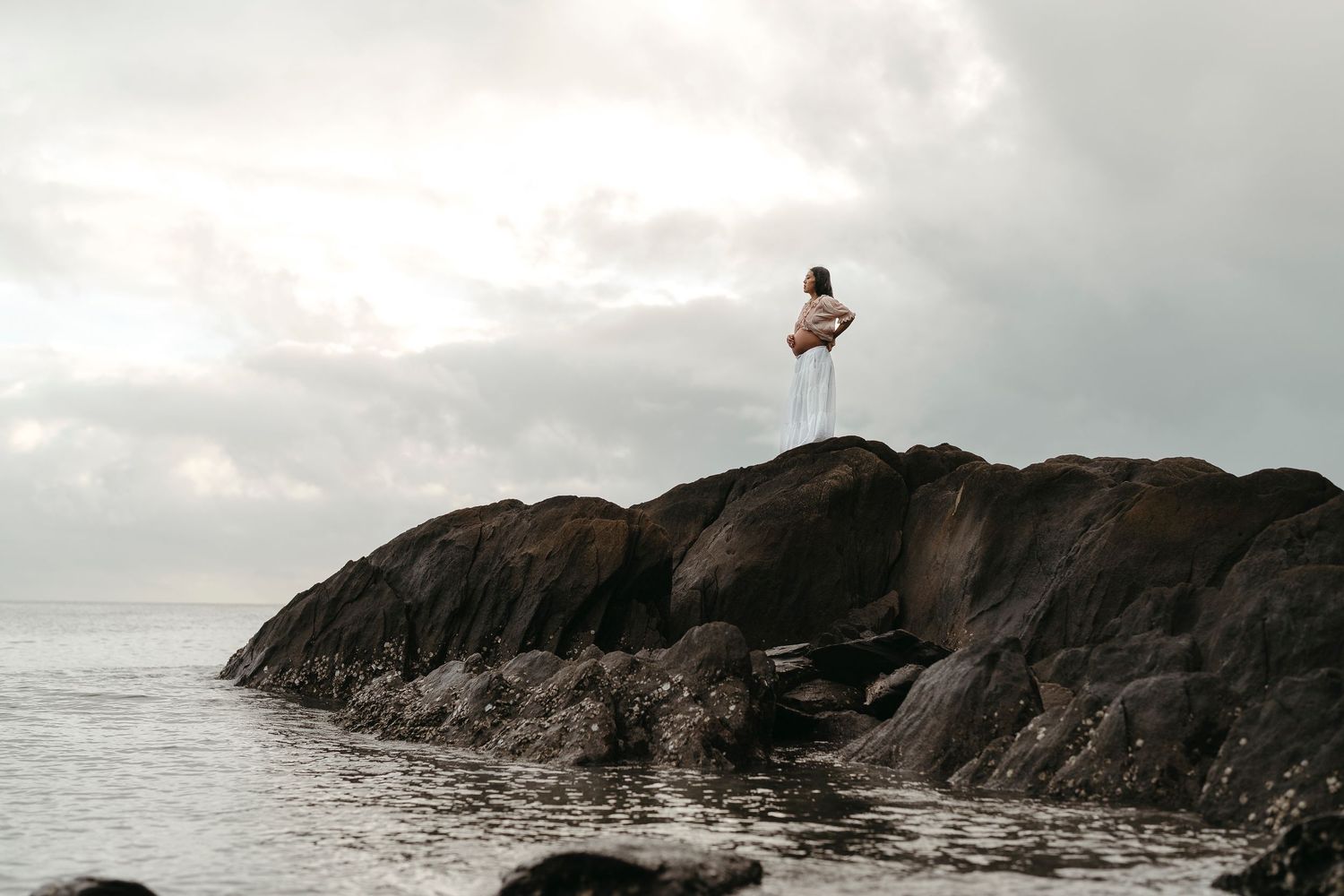 Person standing on rocky coastal outcrop wearing white, gazing at overcast ocean horizon.