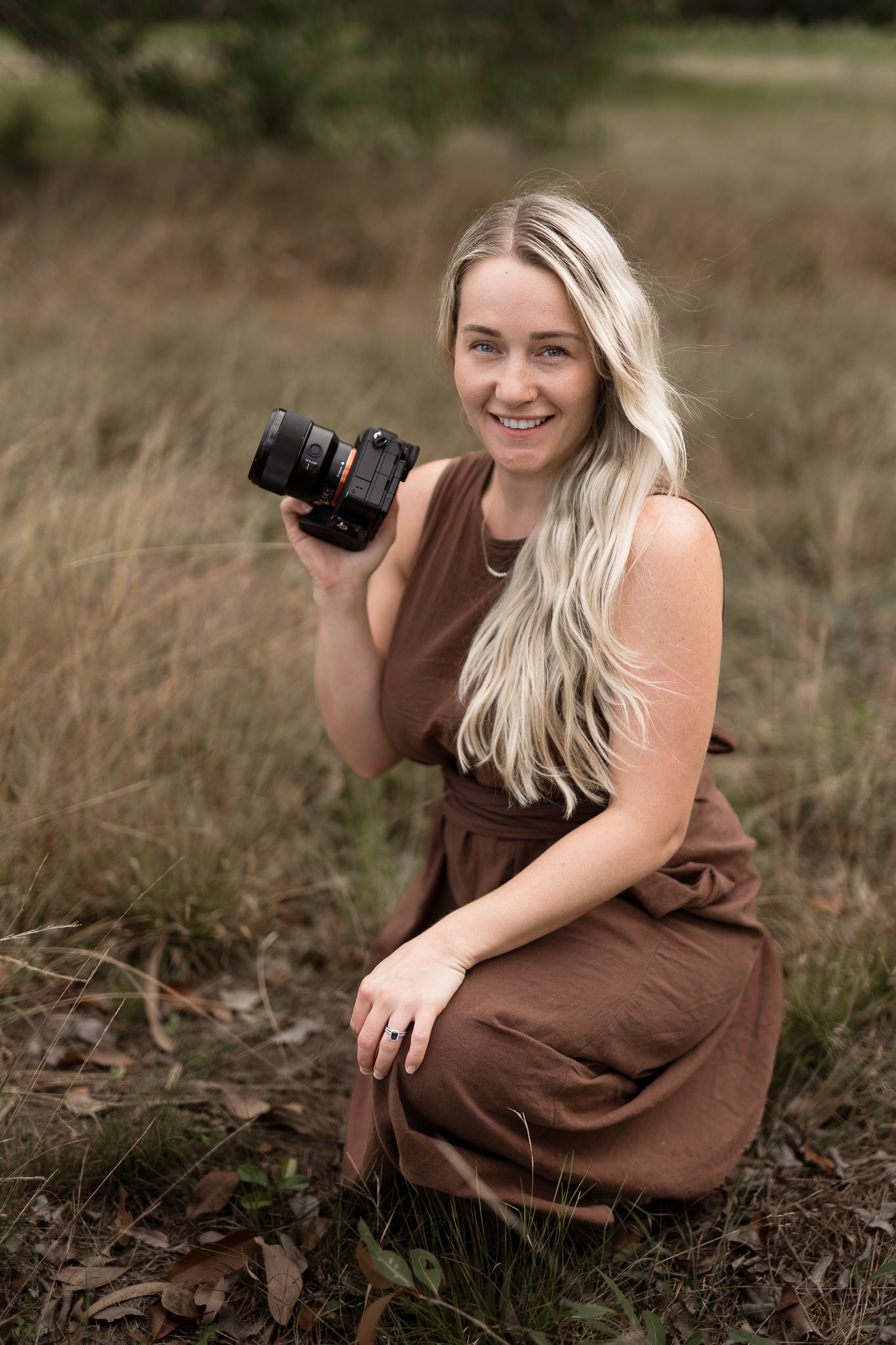 Smiling blonde female photographer kneeling in a field holding a professional camera outdoors.