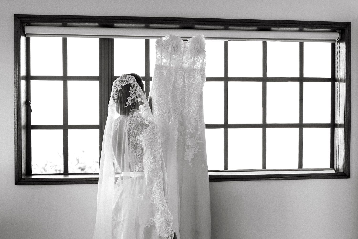 Black and white photo of a lace wedding dress and veil hanging by a window.