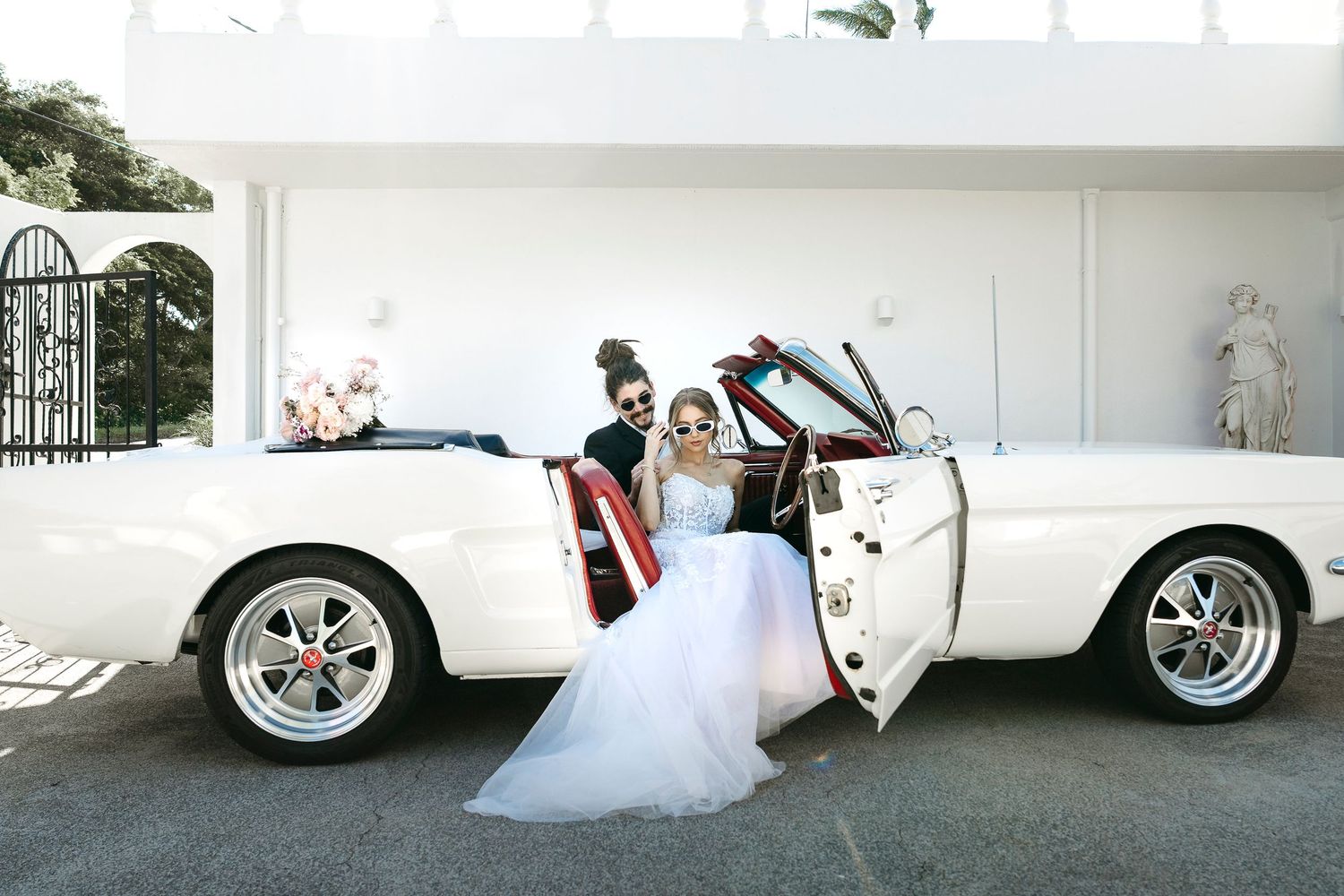 Two brides pose beside a classic white convertible Mustang on their wedding day.