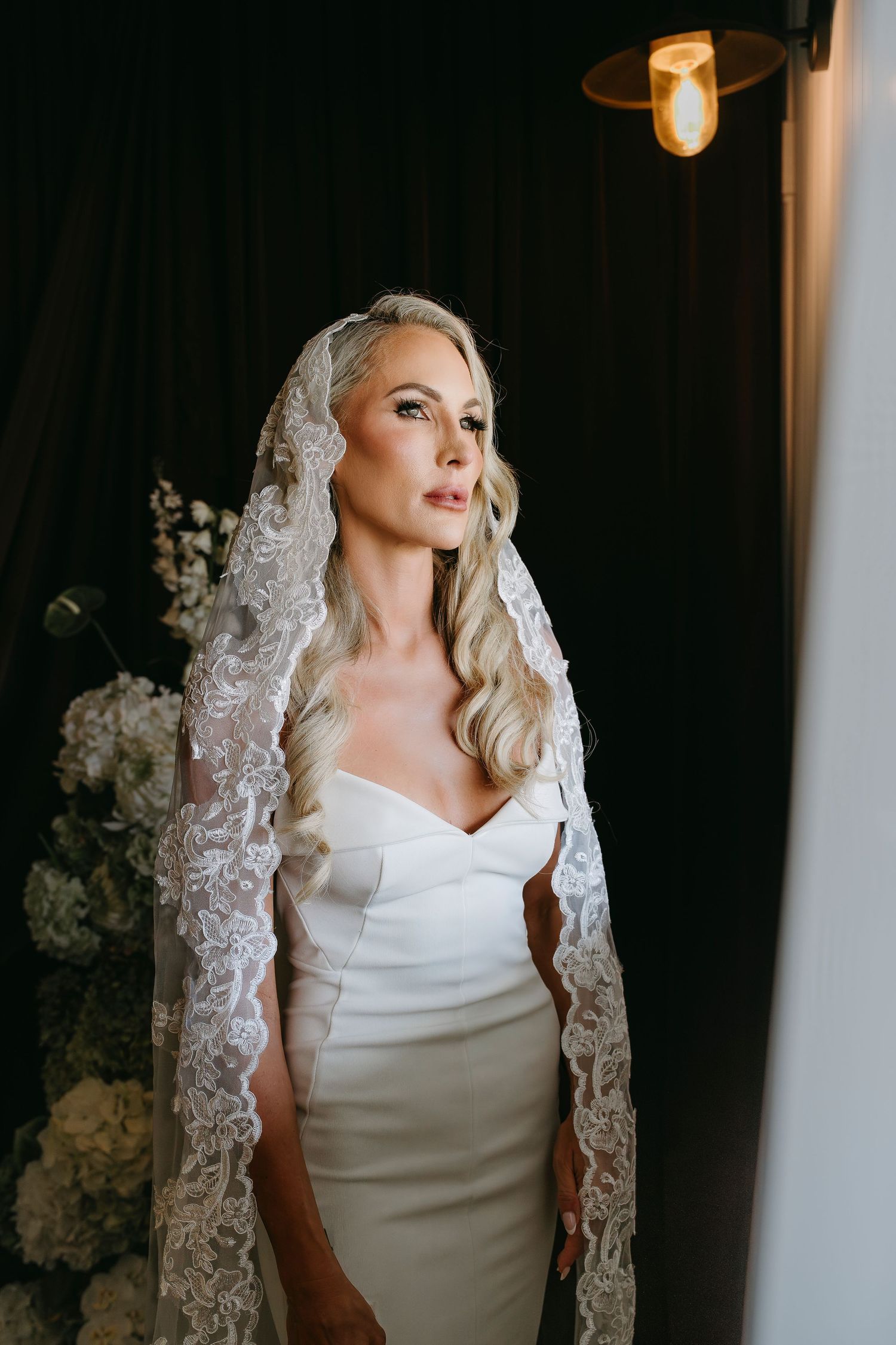 Elegant blonde bride in a strapless gown and lace mantilla veil poses in a softly lit interior setting.