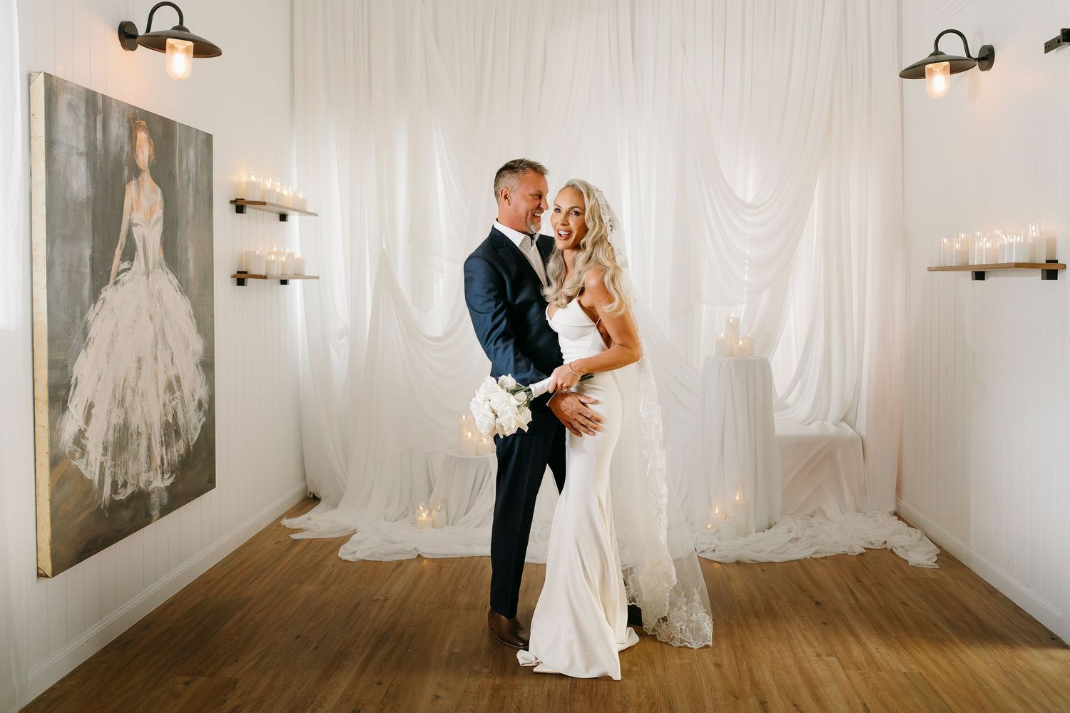 Elegant bride and groom embracing in a white-draped wedding venue with candlelit shelves and a bridal portrait painting.