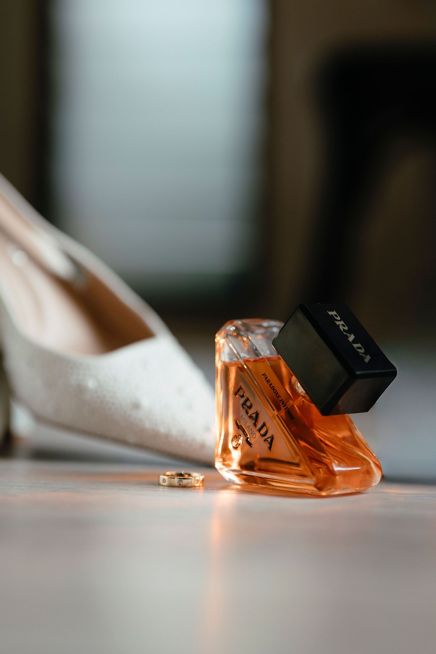 Prada perfume bottle with orange fragrance beside white bridal heels and gold wedding ring on white surface.