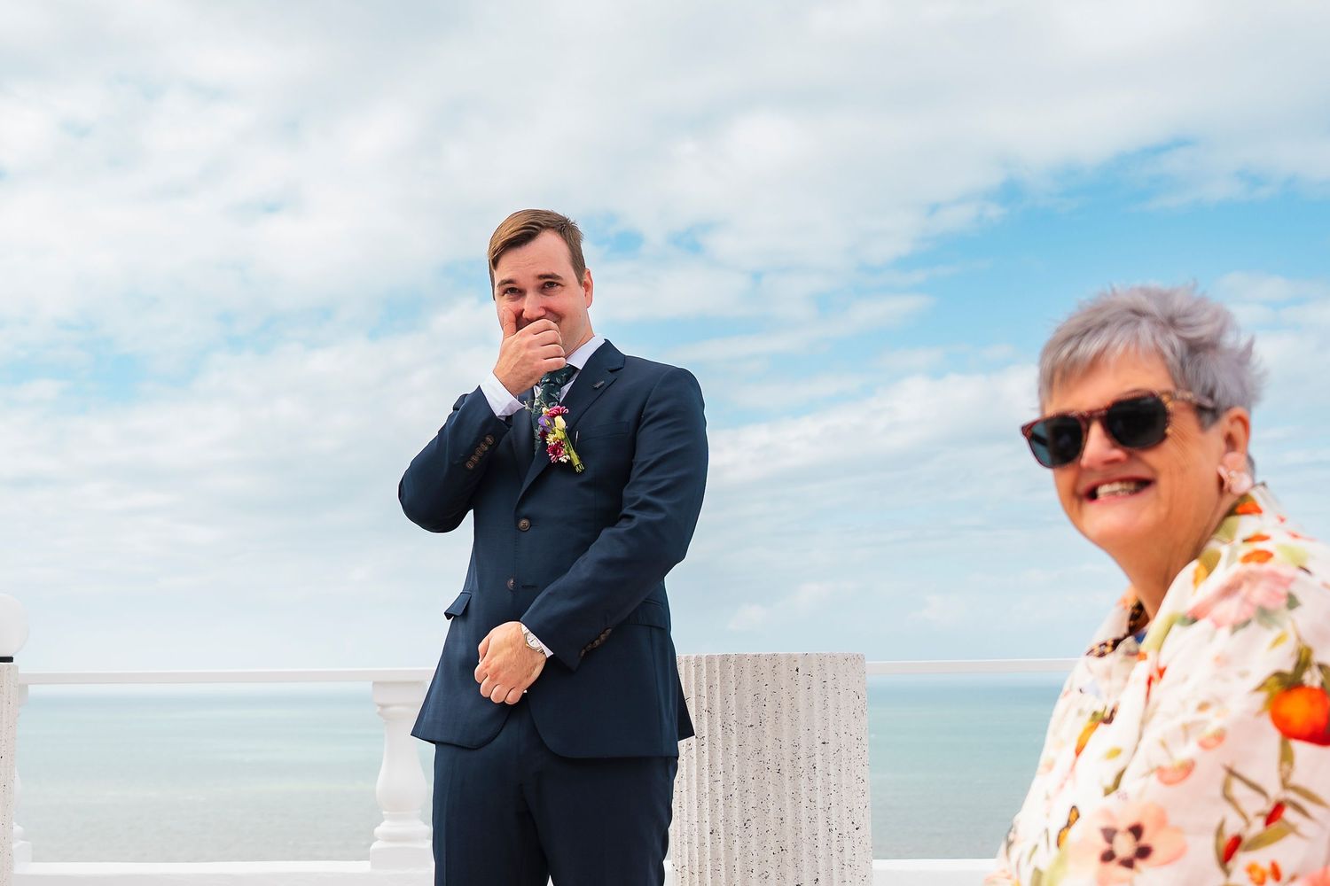 Emotional groom covering his mouth at outdoor beach wedding ceremony, with guest in floral shirt standing nearby.
