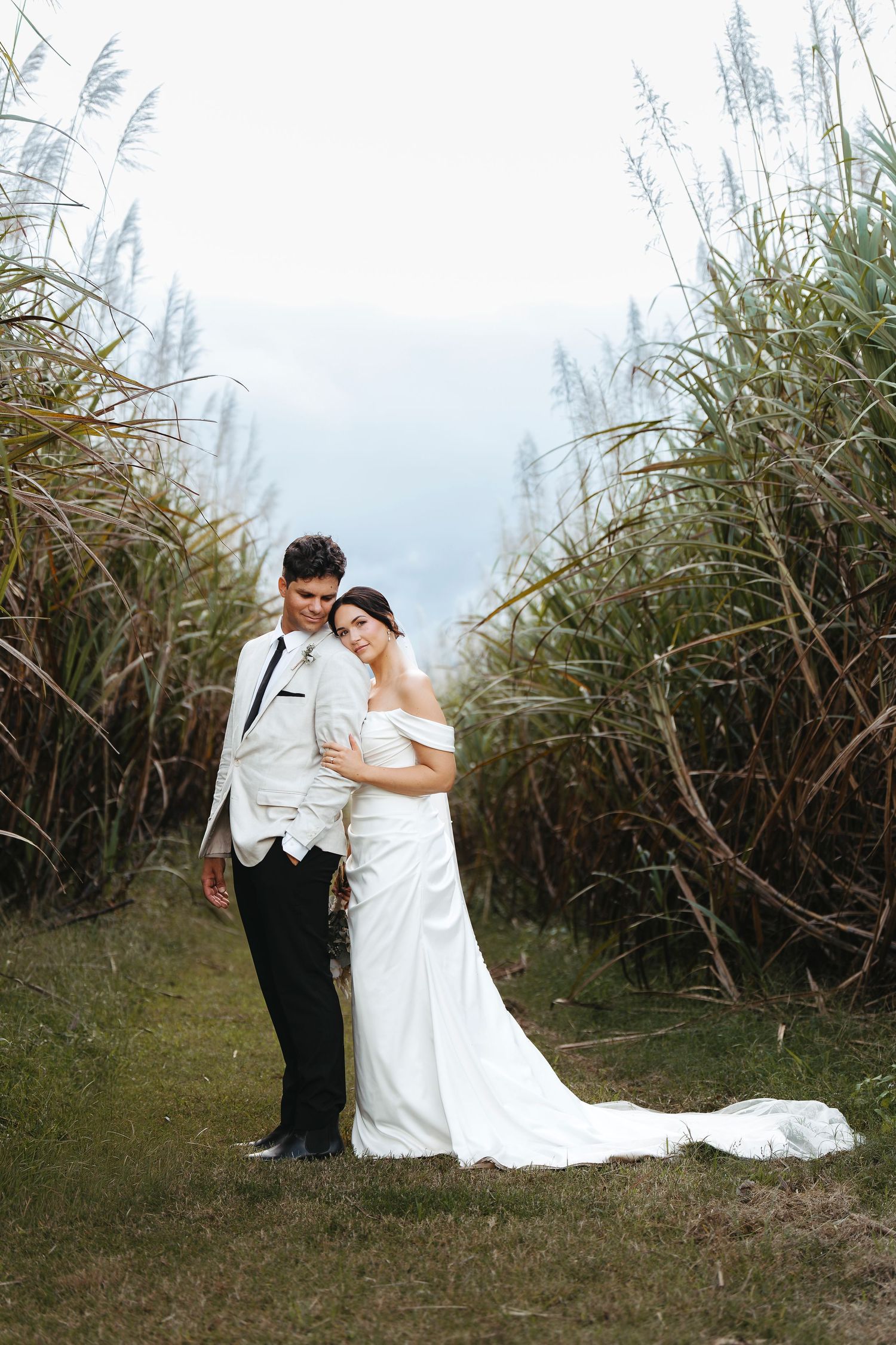 Bride in white off-shoulder gown leans on groom in white suit, posing among tall reeds outdoors.