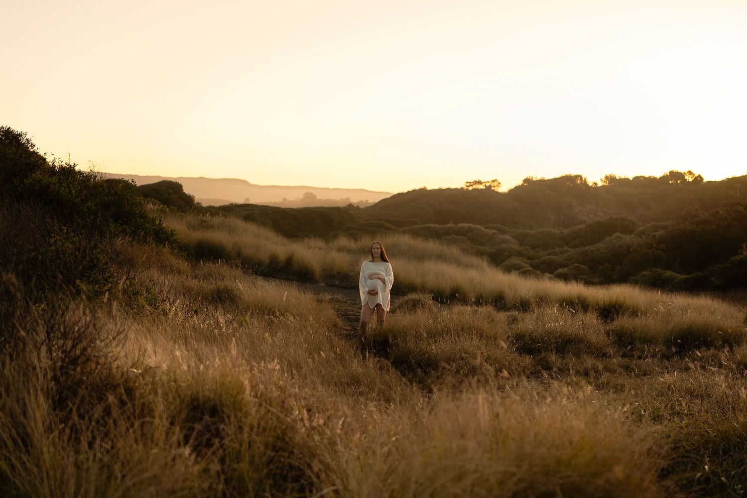 A white dog runs through golden tall grass at sunset in a scenic hilly landscape.