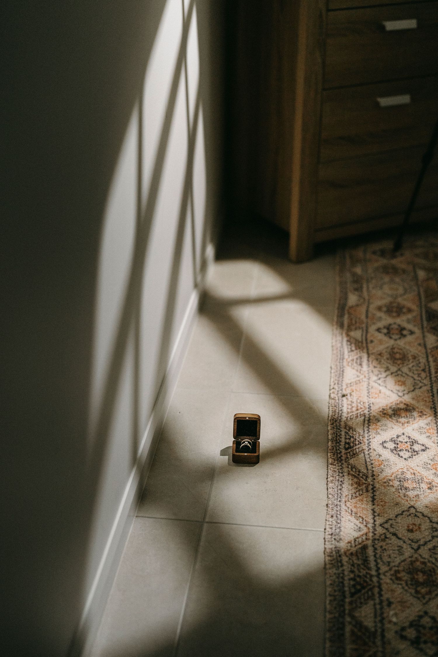 A gold ring in a velvet box sits on sunlit tile floor beside a patterned rug and wooden dresser.